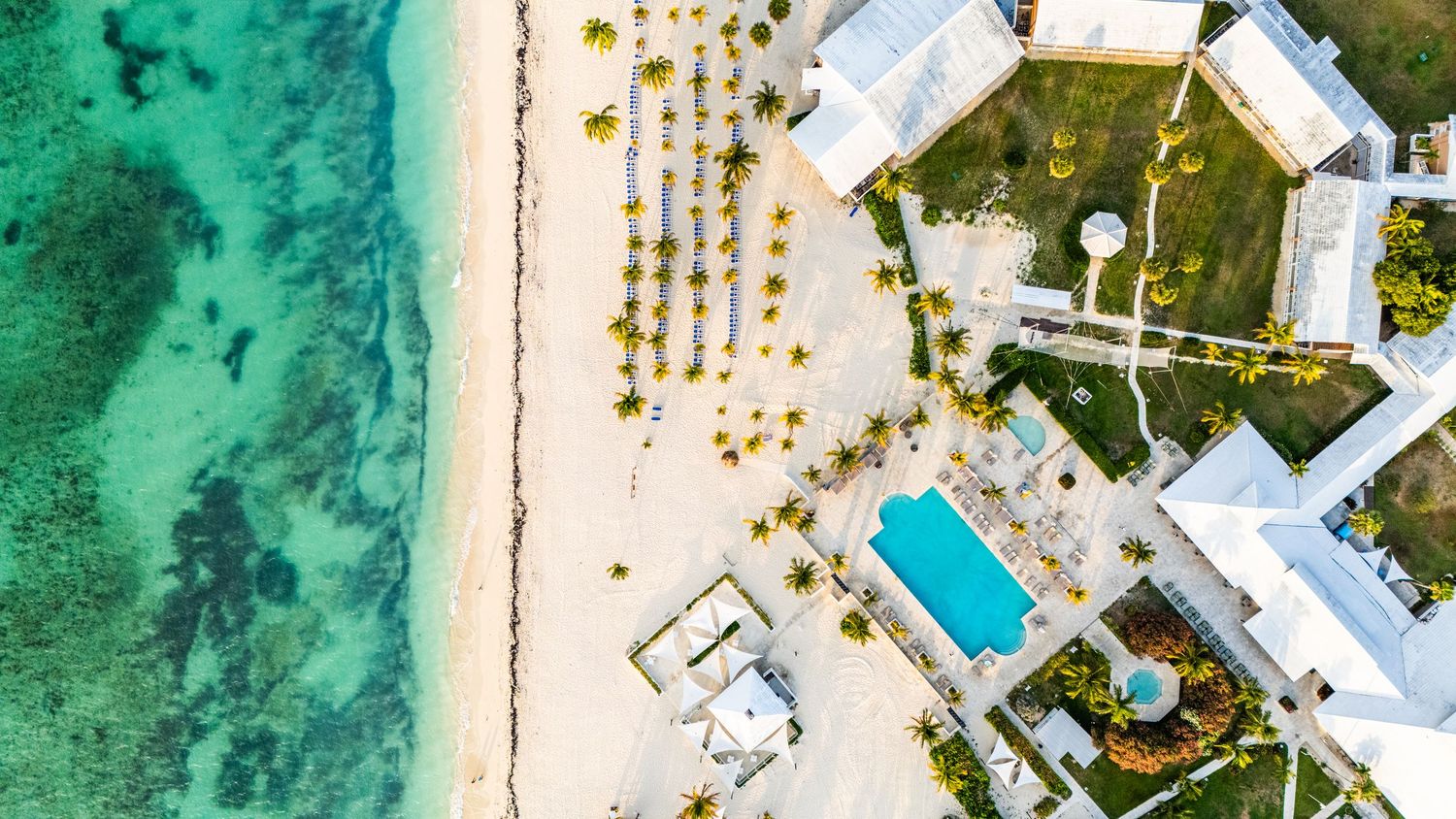 Aerial view of a beachfront resort with swimming pool and turquoise ocean waters along a pristine white sand beach.