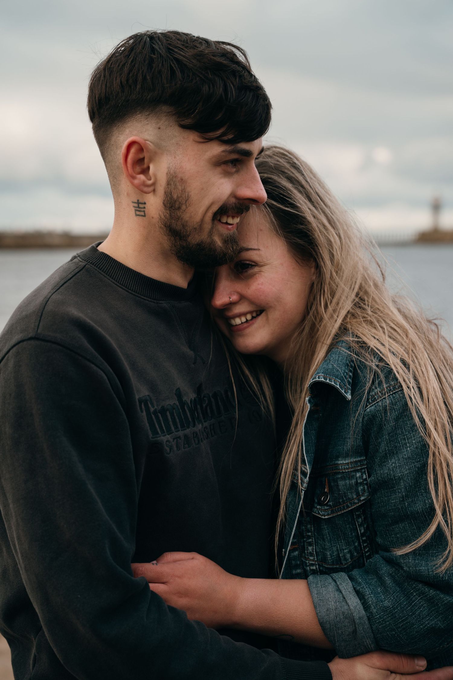 Happy young couple embracing outdoors near water, man with neck tattoo smiling at blonde woman.
