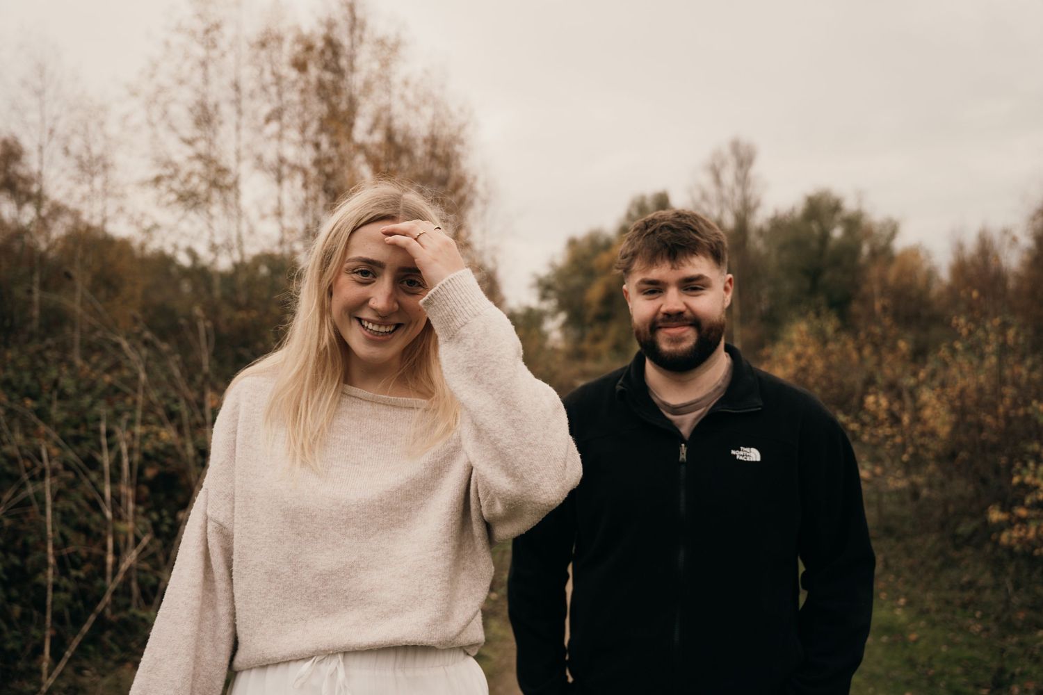 Laughing blonde woman and smiling bearded man standing together in an autumn outdoor setting.