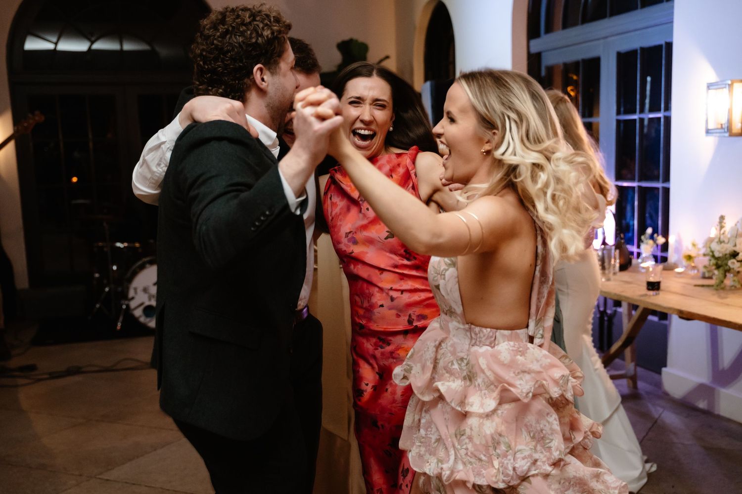 Three joyful wedding guests in formal attire laughing and dancing together at a reception.