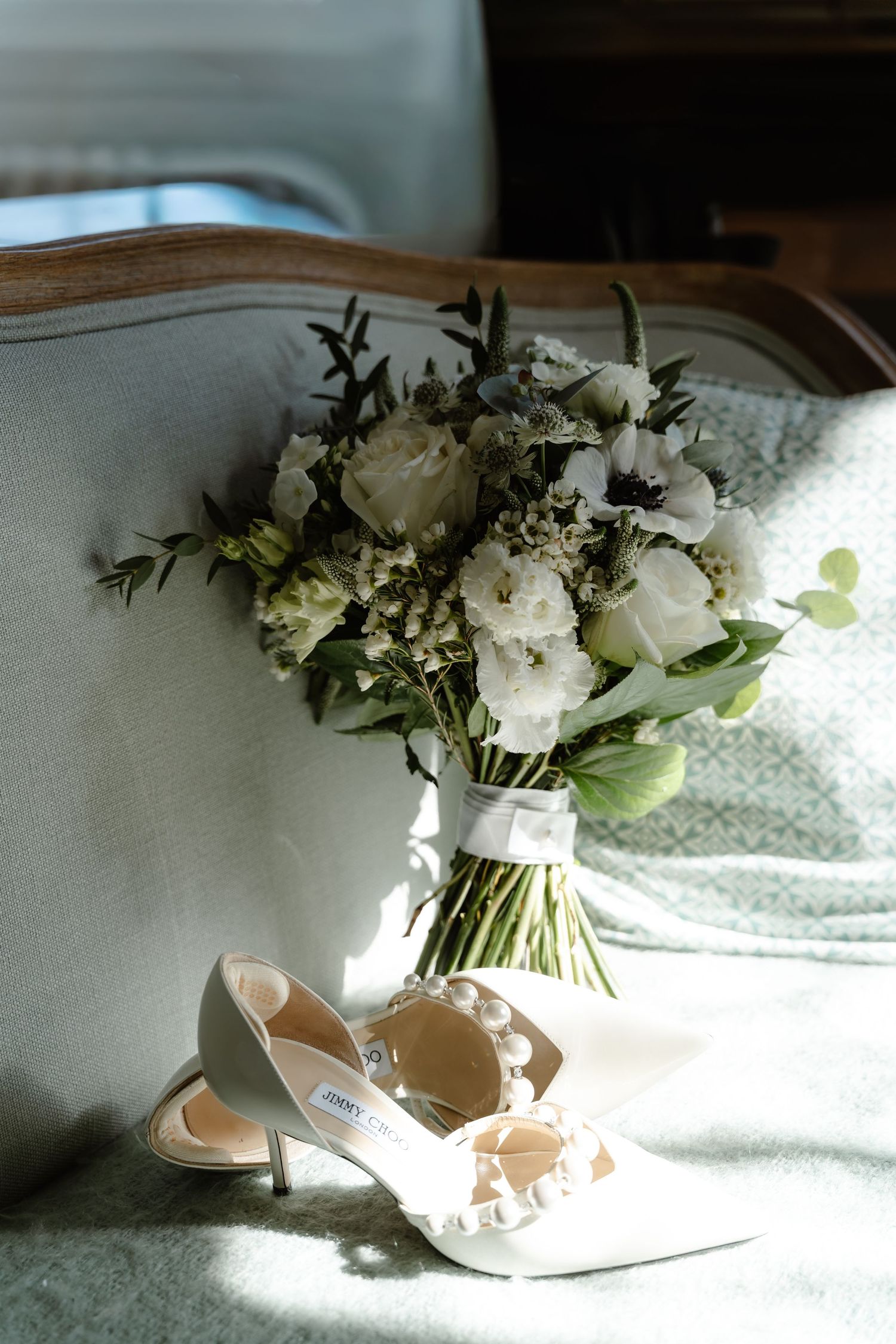 Bridal bouquet of white flowers and greenery beside Jimmy Choo heels on a vintage chair.