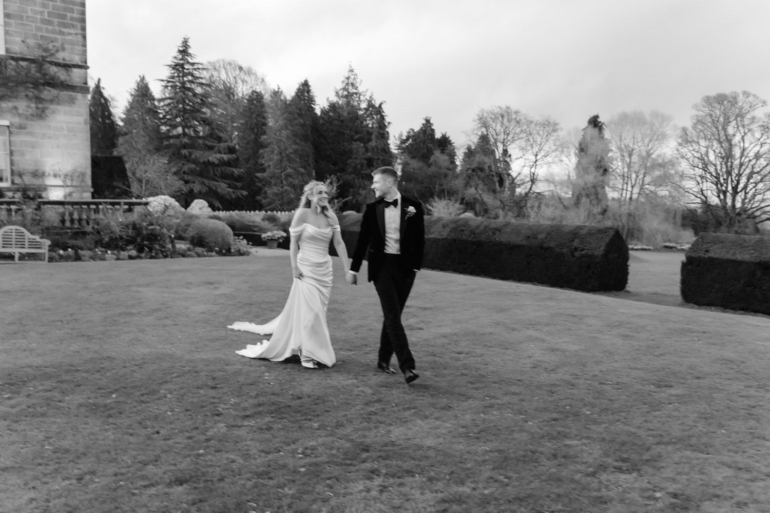 Bride and groom walk across a manicured lawn in black and white, with formal garden hedges and stone architecture behind them.