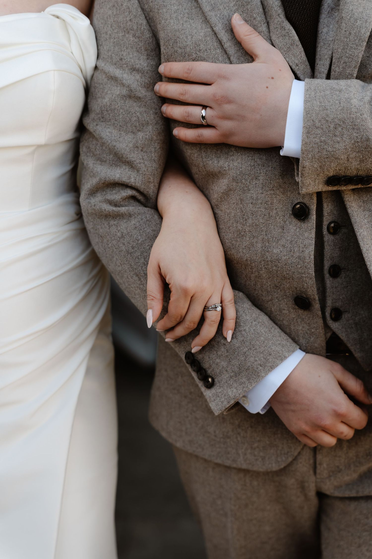 Close-up of bride and groom showing wedding rings while she holds his arm in a grey tweed suit.