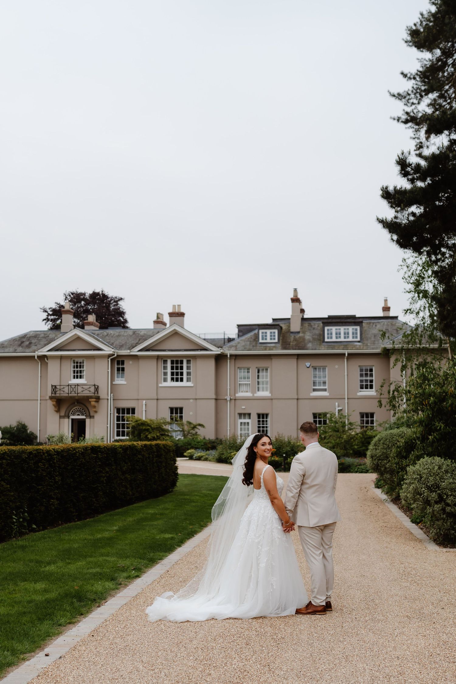 Bride and groom hold hands on a gravel path before a grand Georgian manor house surrounded by manicured hedges.