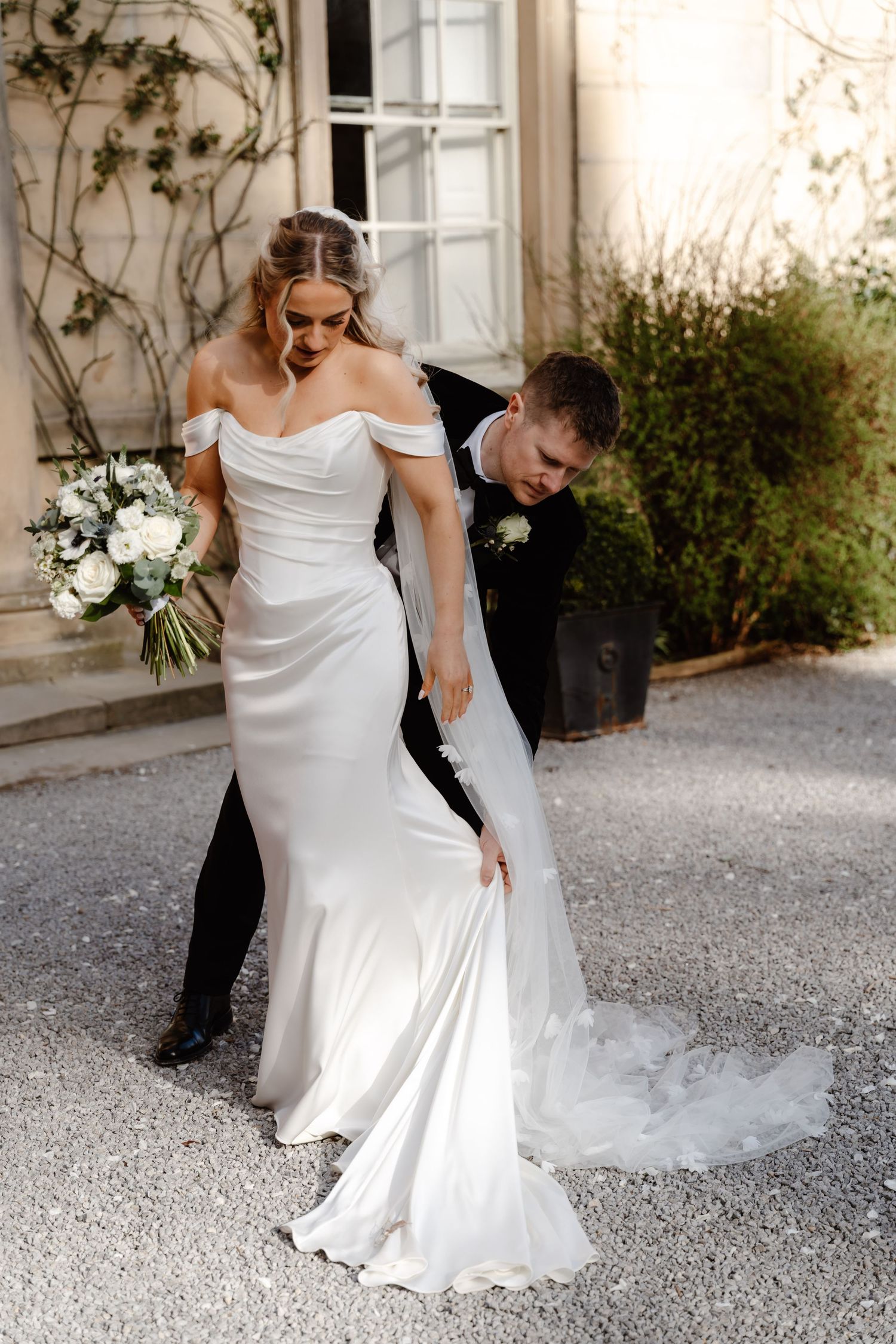 Groom carefully arranging brides off-shoulder wedding dress train outside a charming ivy-covered venue.