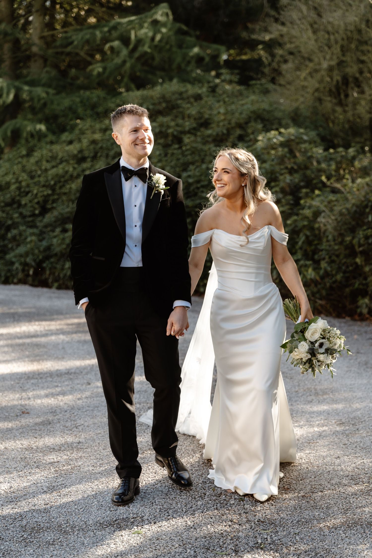 Bride and groom walk hand in hand on gravel path, she in off-shoulder white gown holding bouquet, he in black tuxedo.