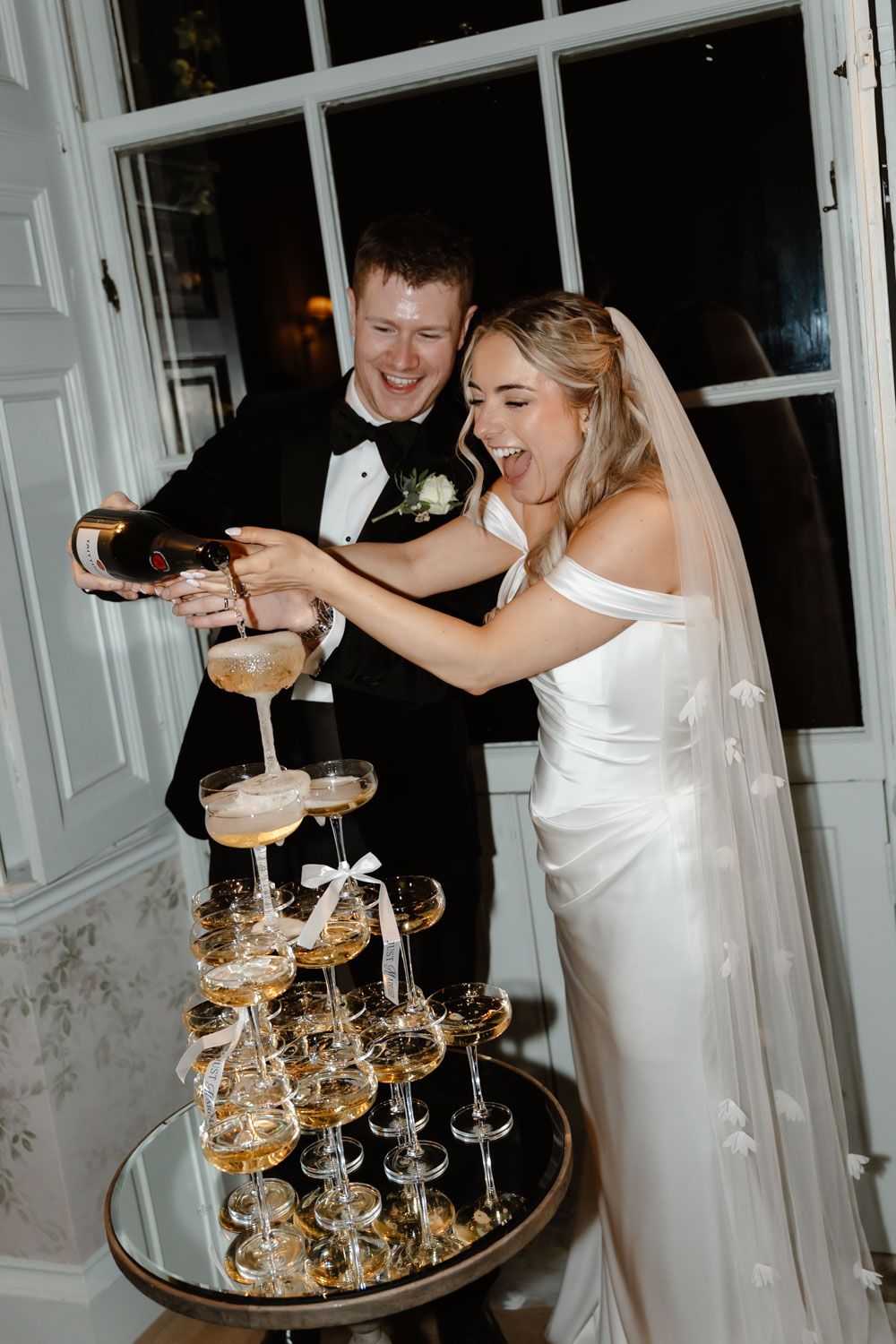 Excited bride and groom pouring champagne tower together at elegant indoor wedding reception celebration.