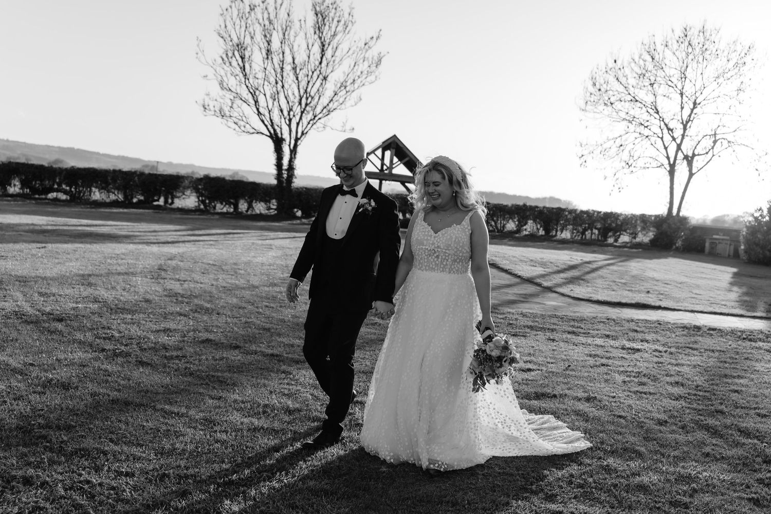 Black and white photo of a bride and groom walking outdoors on their wedding day across a lush green lawn.
