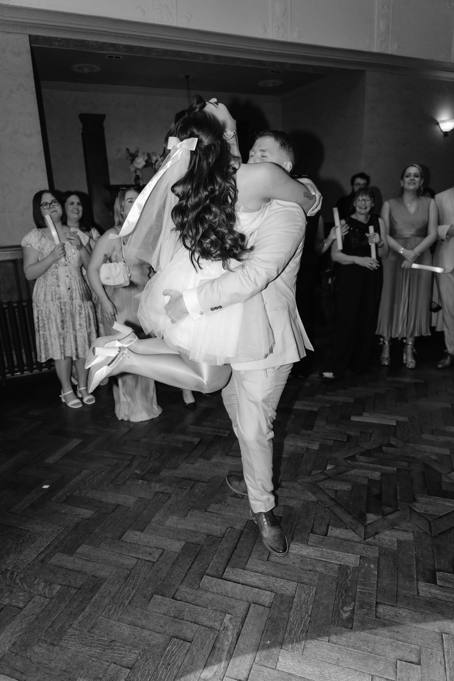 Black and white photo of groom lifting bride during first dance at wedding reception with guests watching.