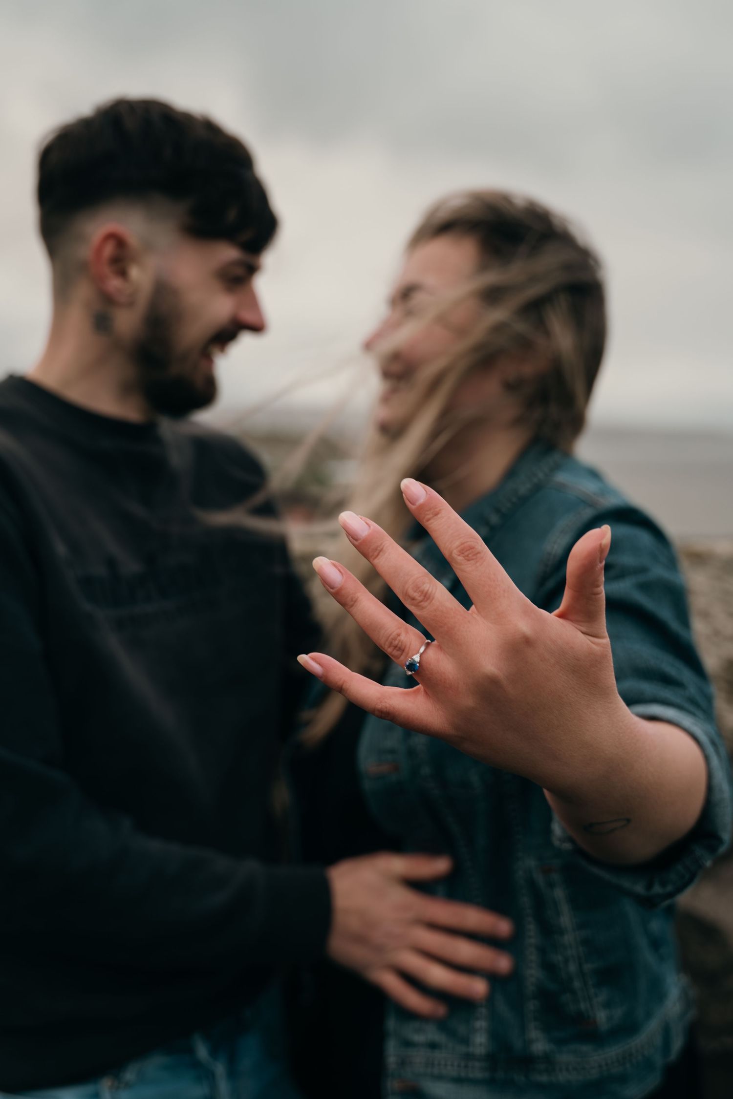 Engaged couple smiling outdoors as woman displays her ring with a dark gemstone toward the camera.