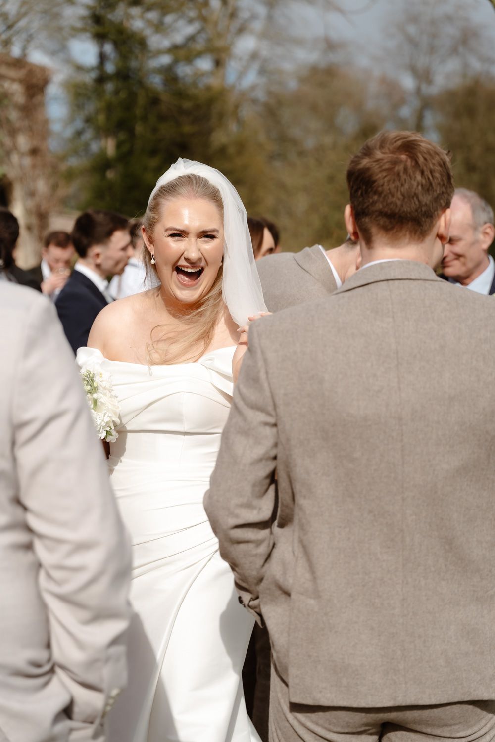 Joyful bride in white off-shoulder gown laughing with groom surrounded by wedding guests outdoors.