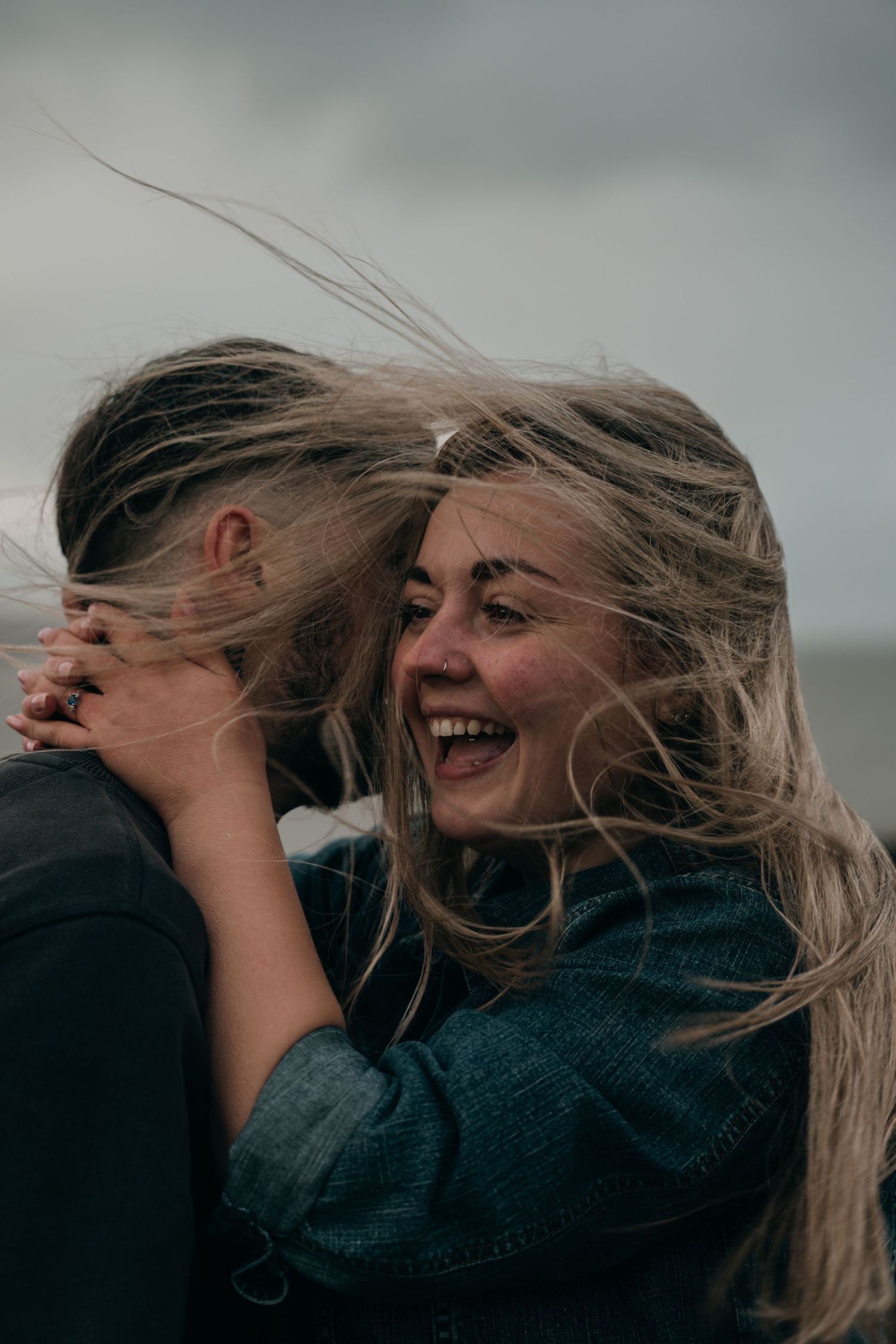 Laughing woman with windswept blonde hair embracing partner outdoors on a cloudy, breezy day.