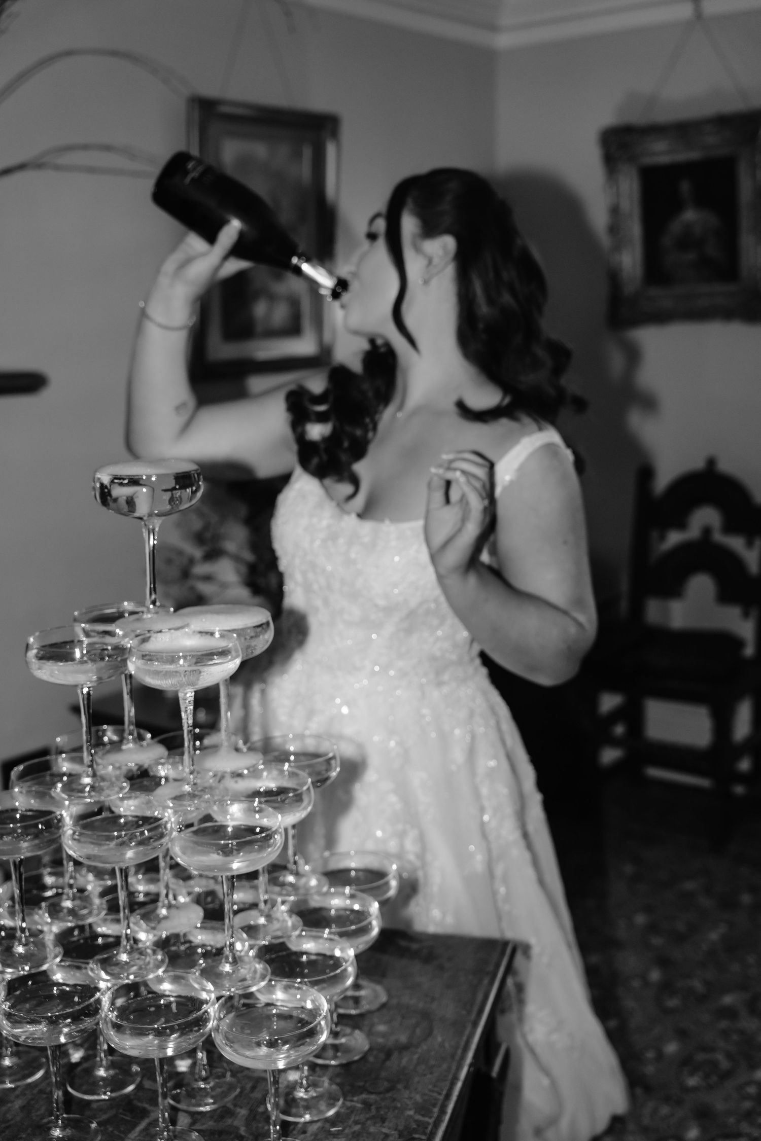 Bride in white gown drinks from champagne bottle beside elegant tower of coupe glasses at wedding reception.