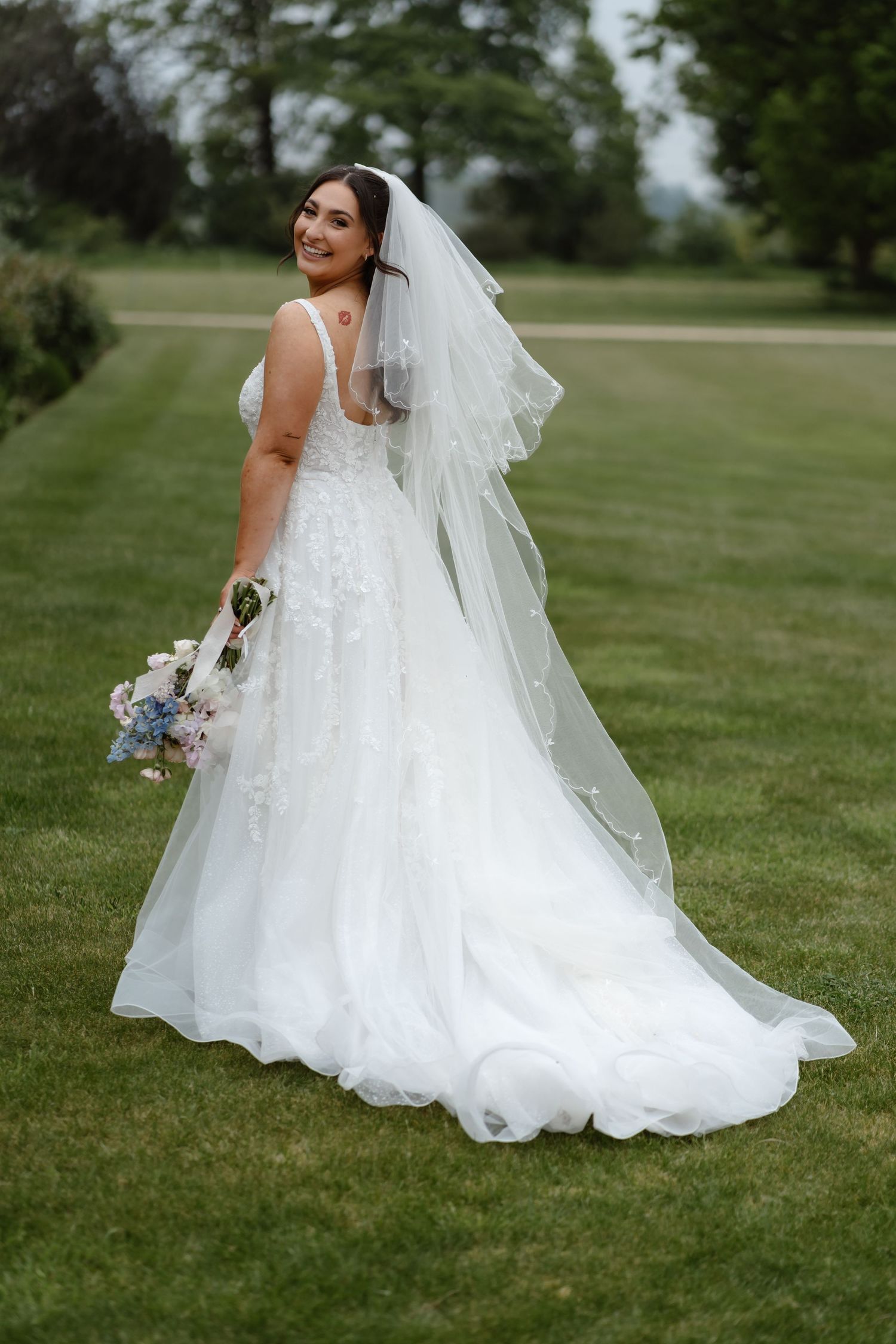 Bride in white floral A-line gown with veil holds bouquet, posing on green lawn at outdoor wedding venue.