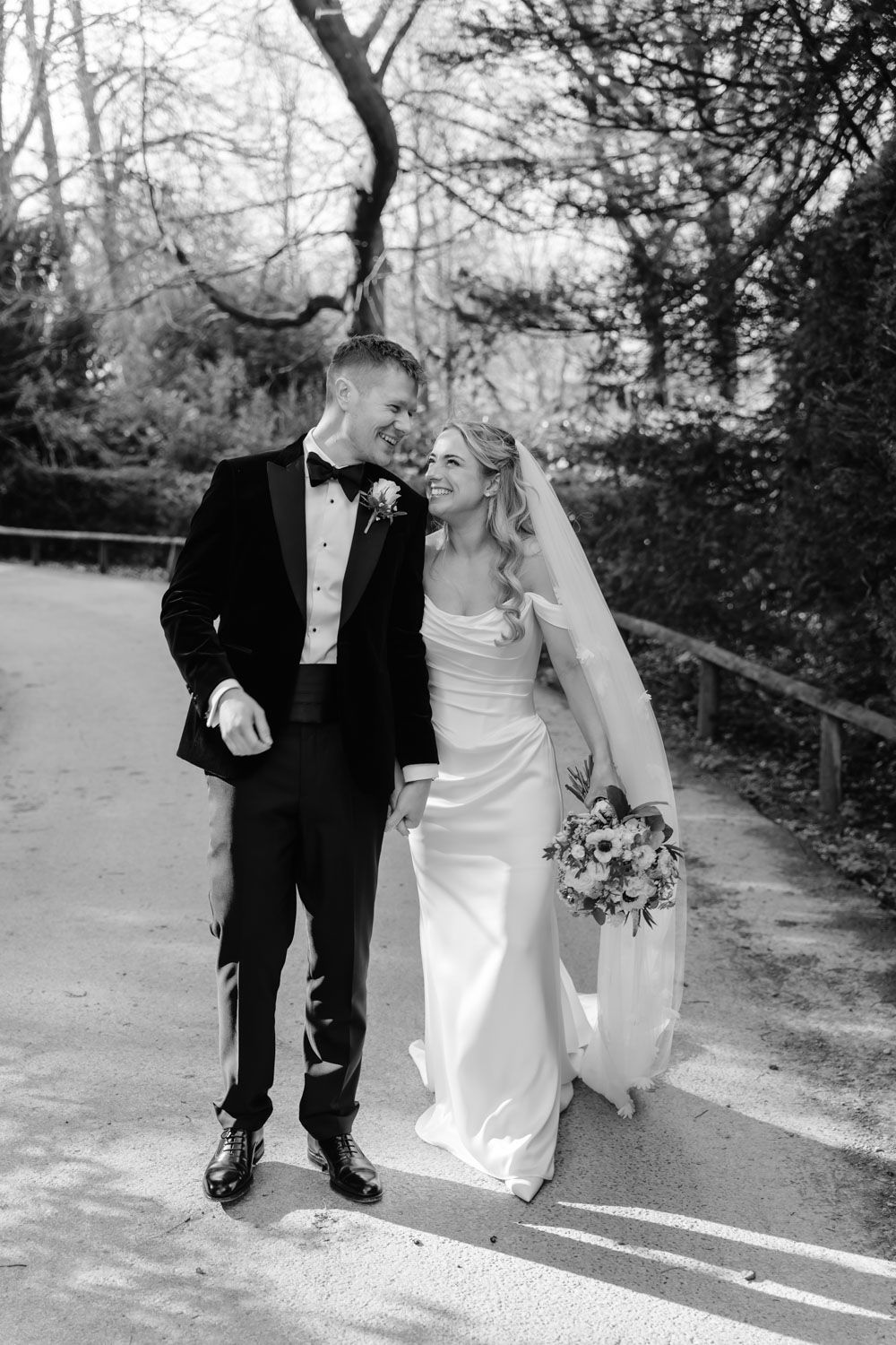 Black and white photo of smiling bride and groom walking together on a sunlit garden path.
