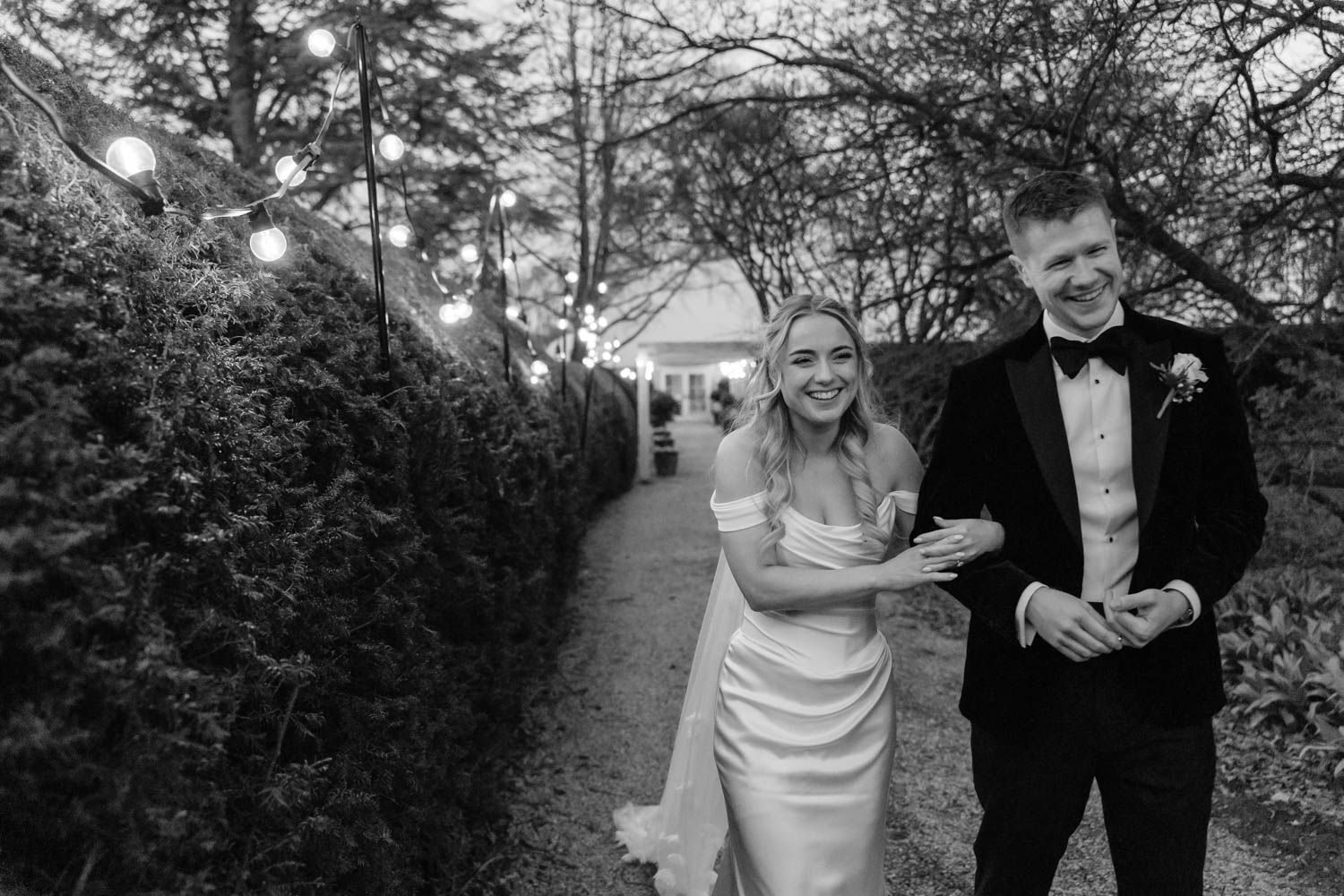 Bride and groom walking together along garden path with fairy lights in black and white.