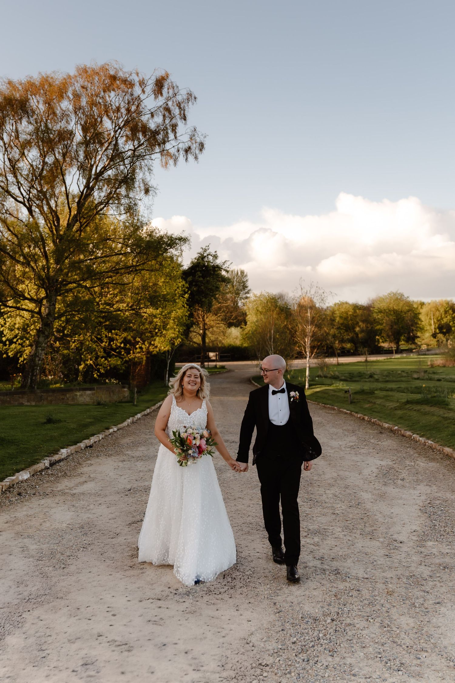 Bride and groom walk hand-in-hand down a gravel path surrounded by lush trees at golden hour.