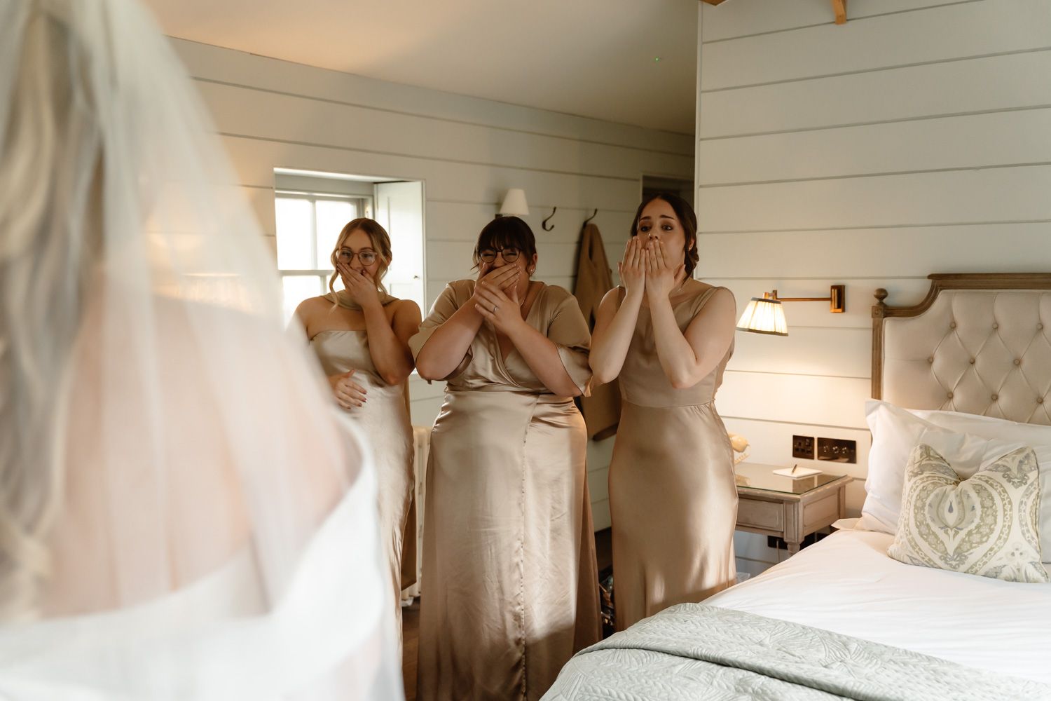 Three bridesmaids in gold satin dresses reacting with awe as bride reveals her wedding gown.