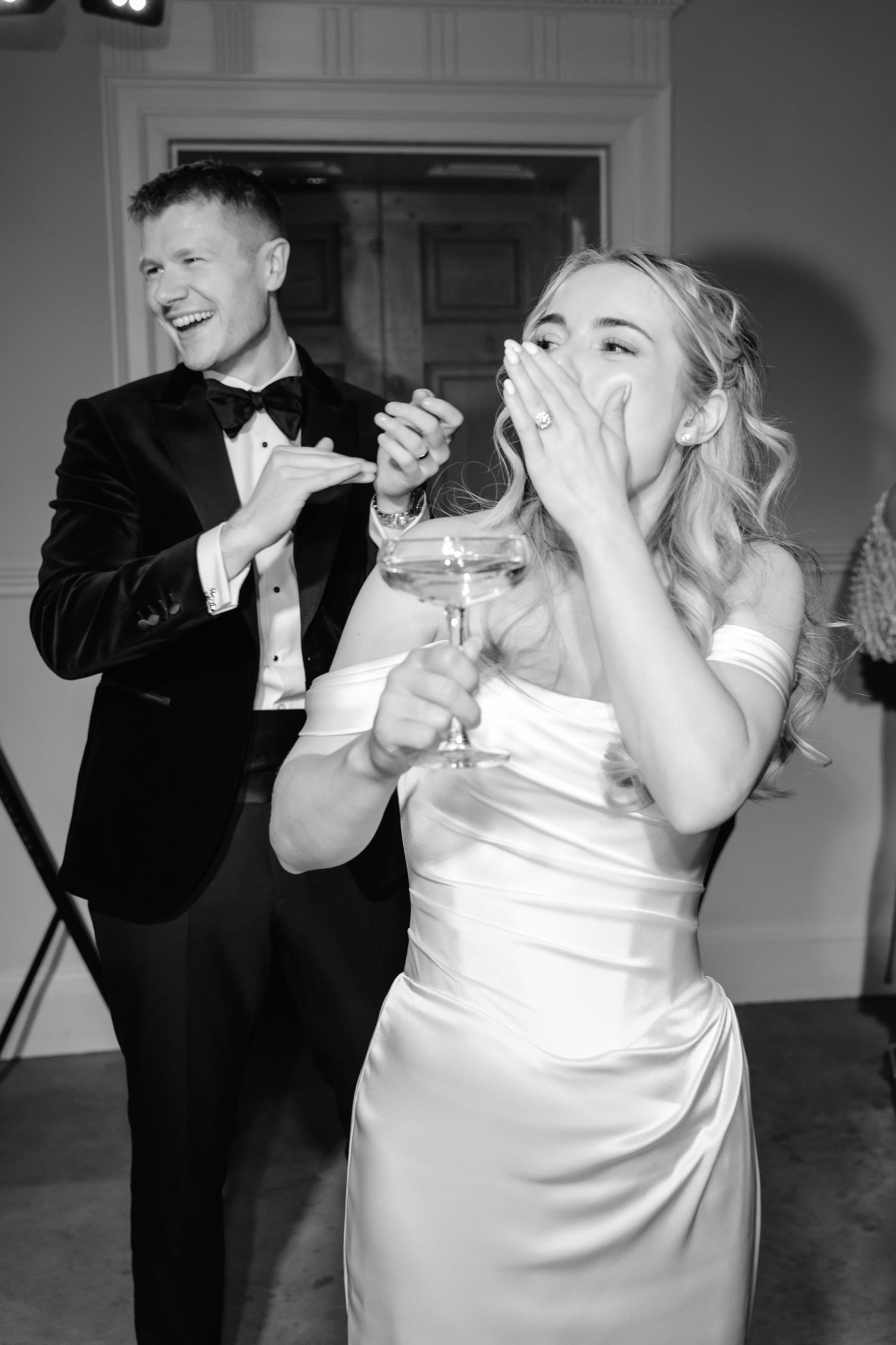 Black and white photo of bride covering her mouth in surprise while groom laughs joyfully behind her at reception.