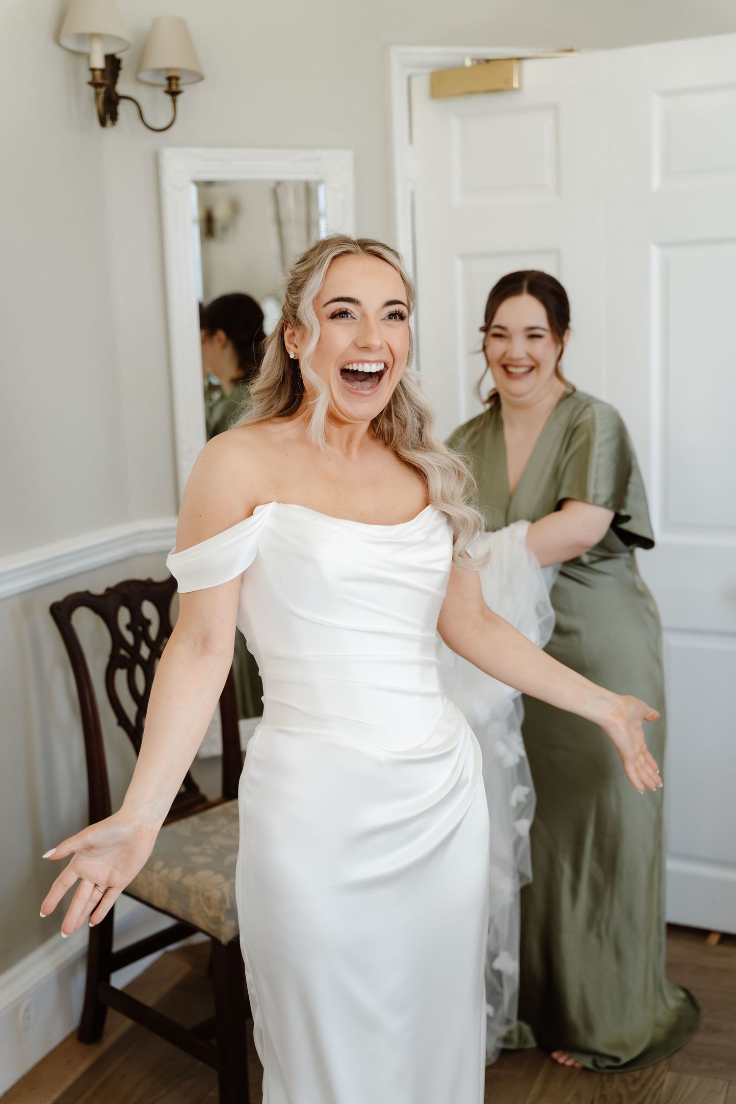 Excited bride in off-shoulder white gown laughing while bridesmaid helps with dress.