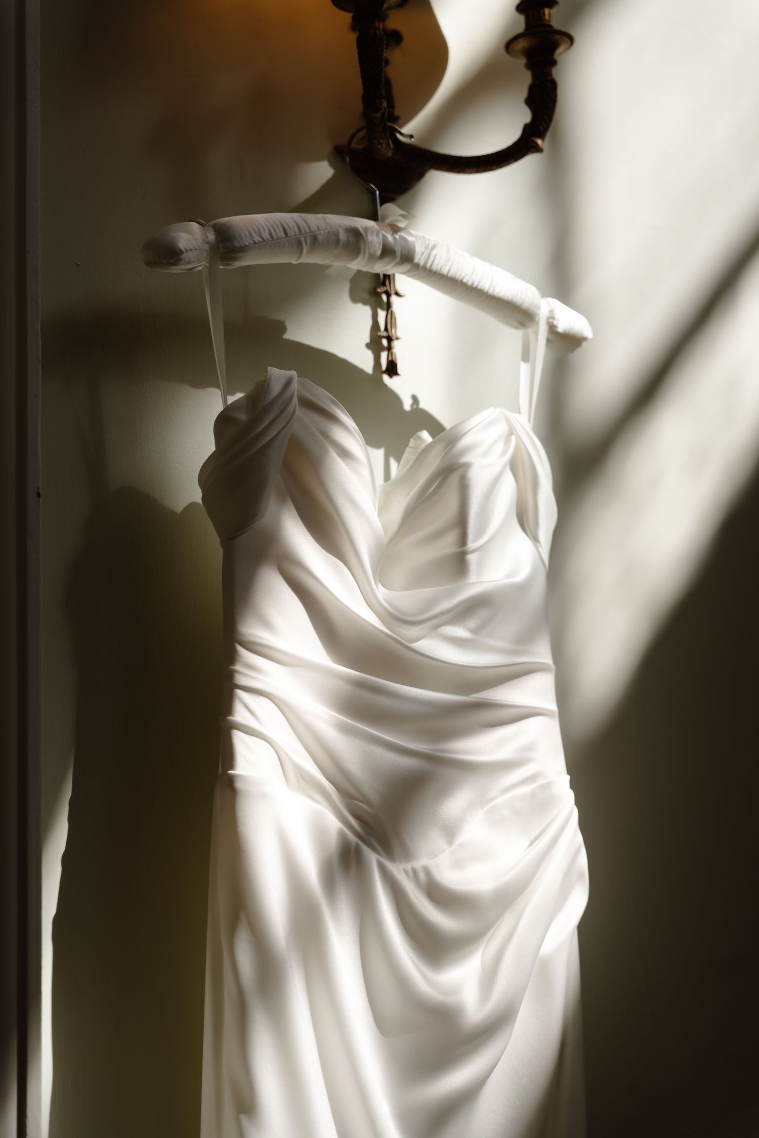 Elegant white satin wedding dress hanging on ornate wall sconce in dramatic natural light.