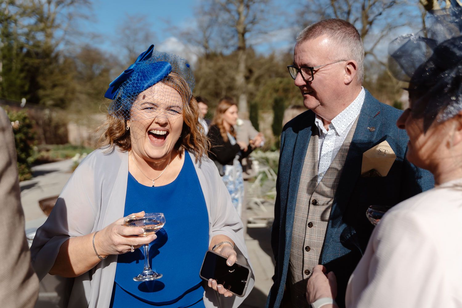 Laughing woman in blue dress and fascinator enjoying drinks with guests at outdoor wedding reception.