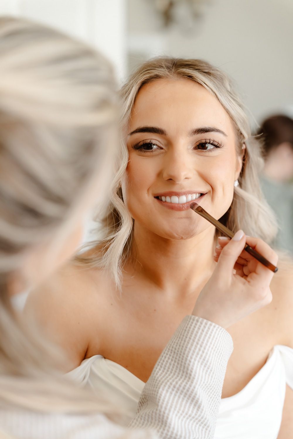 Smiling bride having makeup applied with brush by artist during wedding morning preparations.