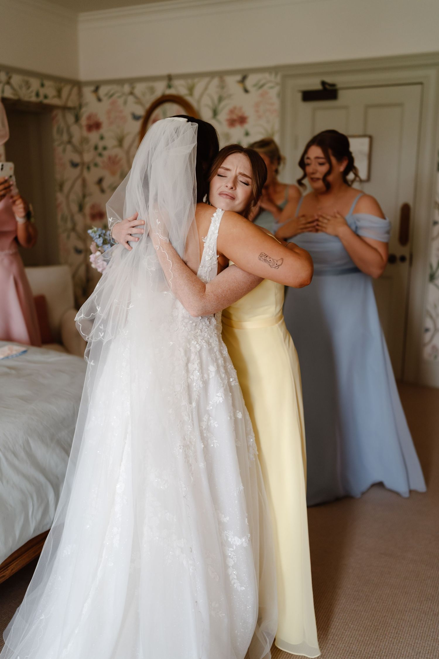 Bride in lace gown and veil shares emotional hug with bridesmaid in yellow dress, while others in blue look on.