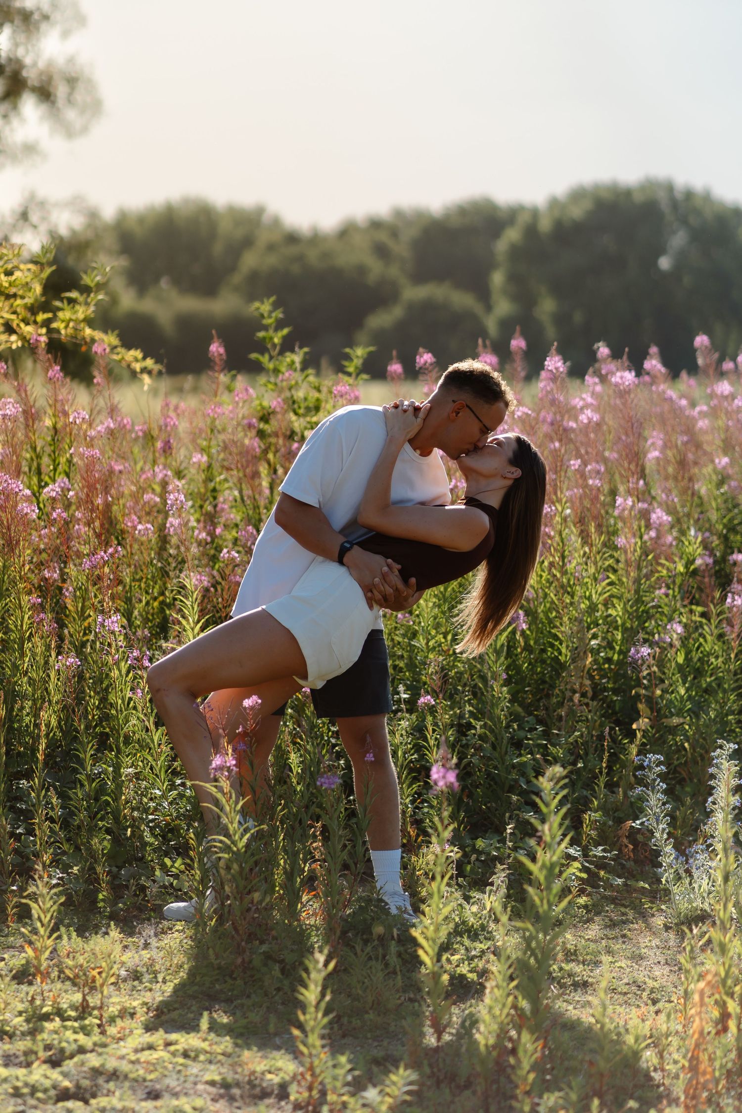 Romantic couple kissing in a blooming pink wildflower field during golden hour sunset.