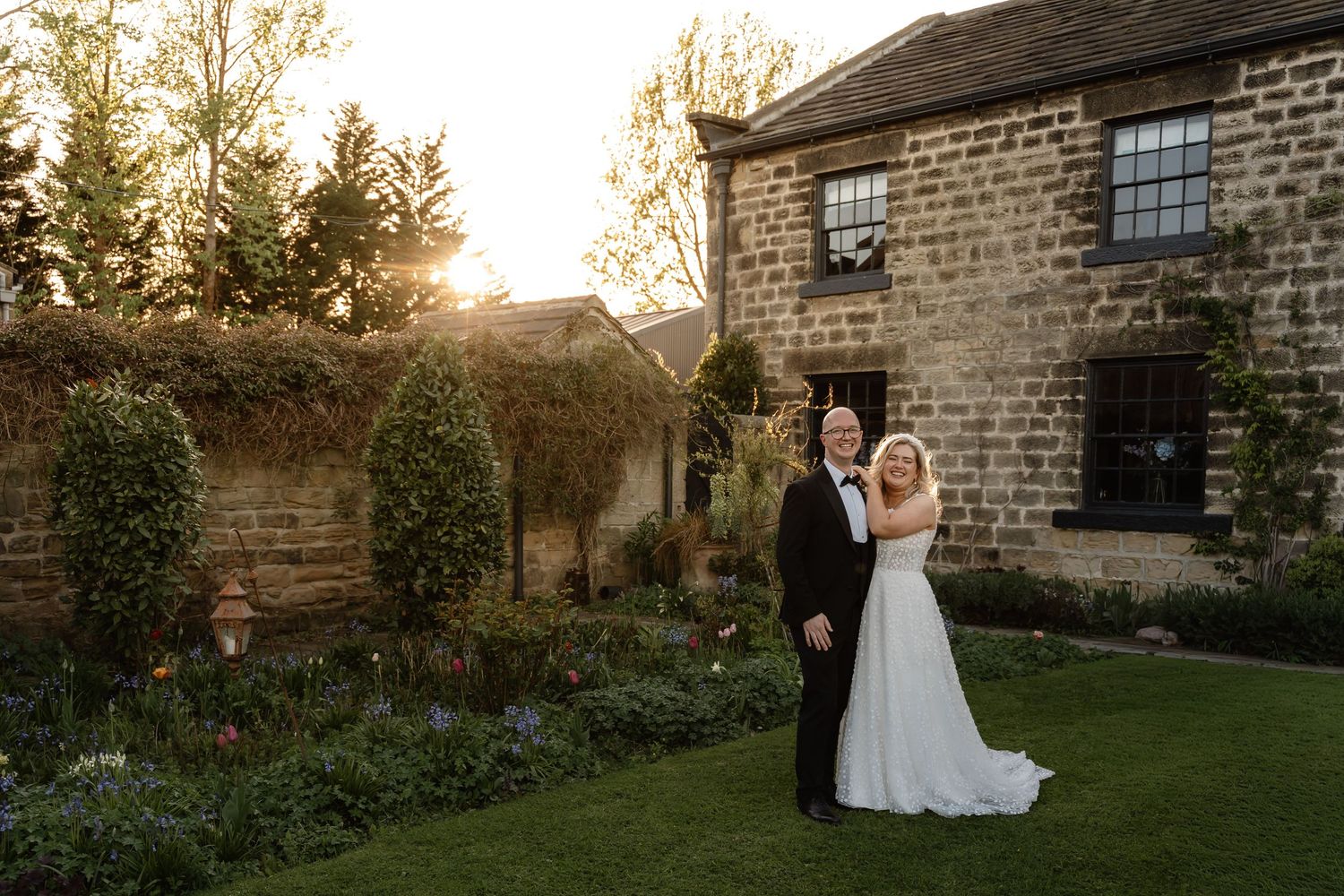 Bride and groom pose in a sunlit garden beside a stone country house at golden hour during their wedding.
