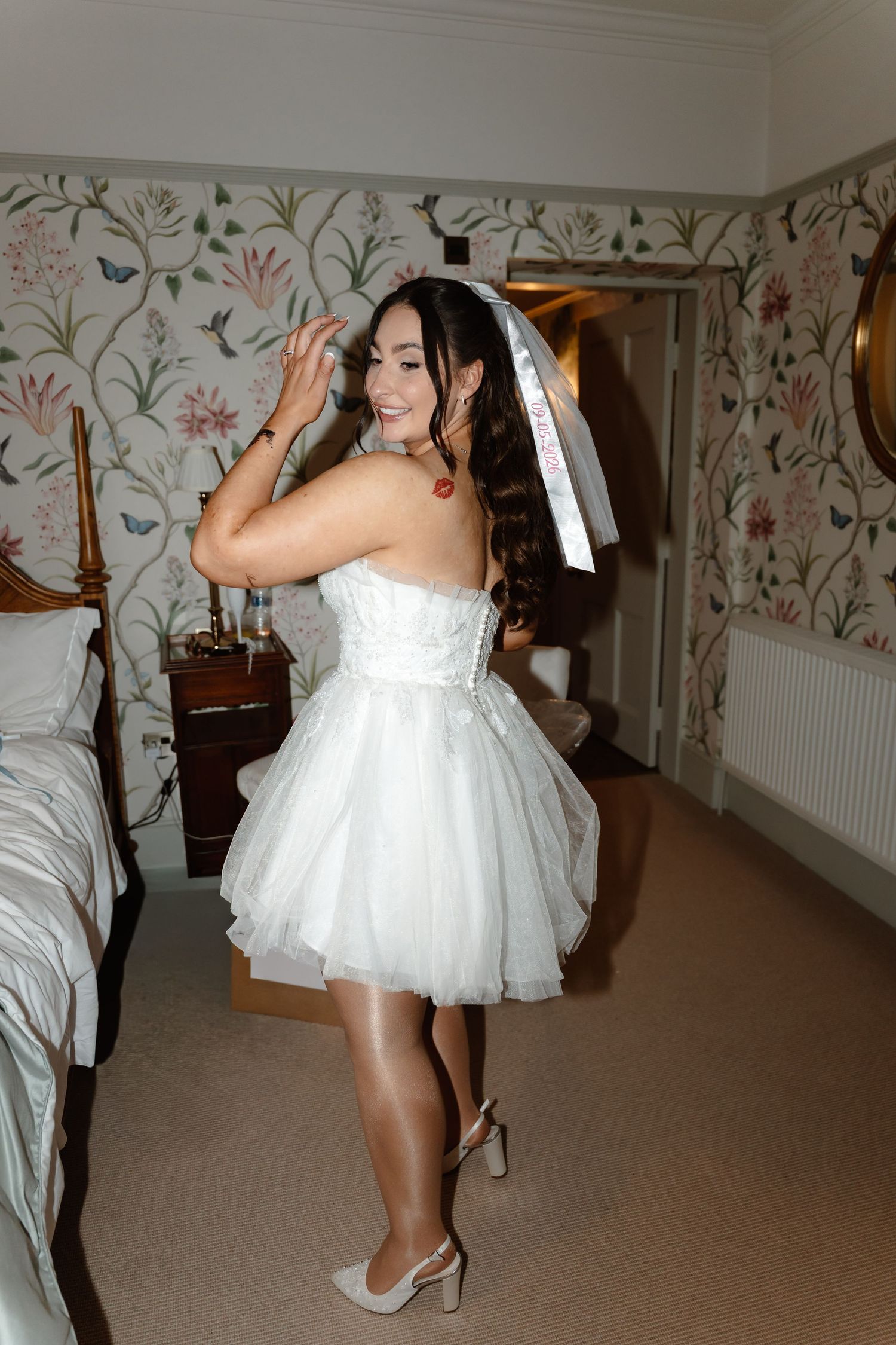 Bride in short white tulle dress and veil poses happily in floral wallpapered bedroom with heels.