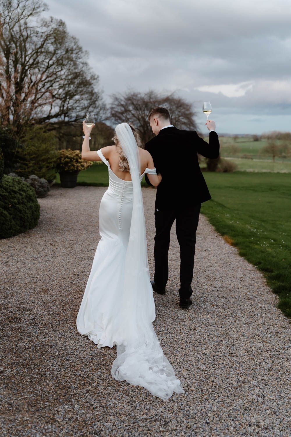 Bride and groom raise champagne glasses while walking away down a gravel path, bride wearing a long veil and fitted gown.