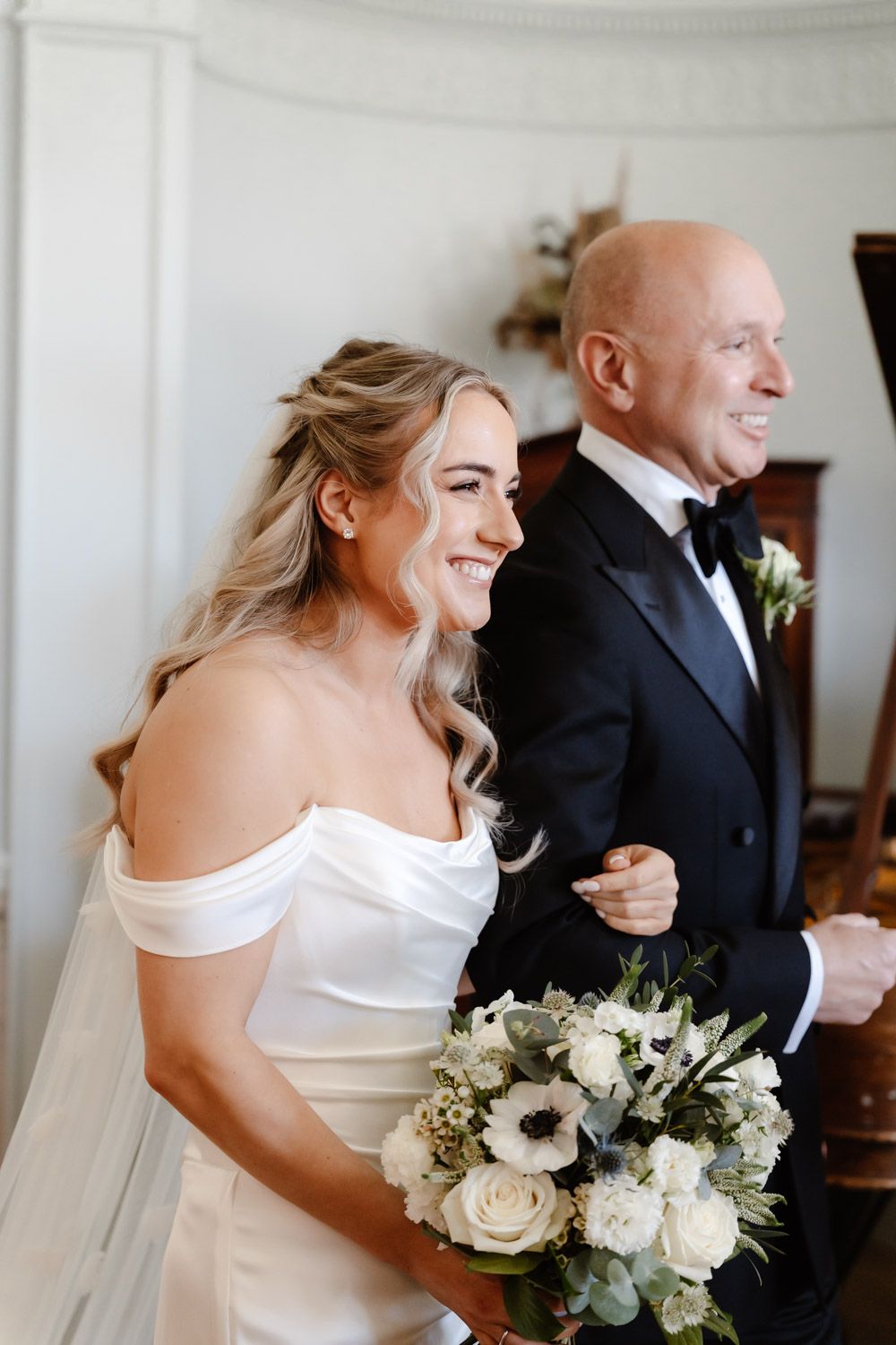 Radiant bride in off-shoulder gown holding white floral bouquet walking down aisle with father.