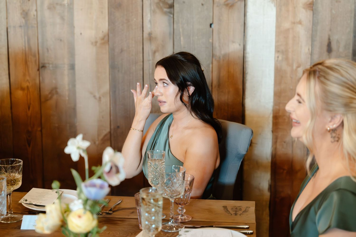 Two women in elegant green dresses seated at a decorated dining table, engaged in lively conversation at a wedding reception.