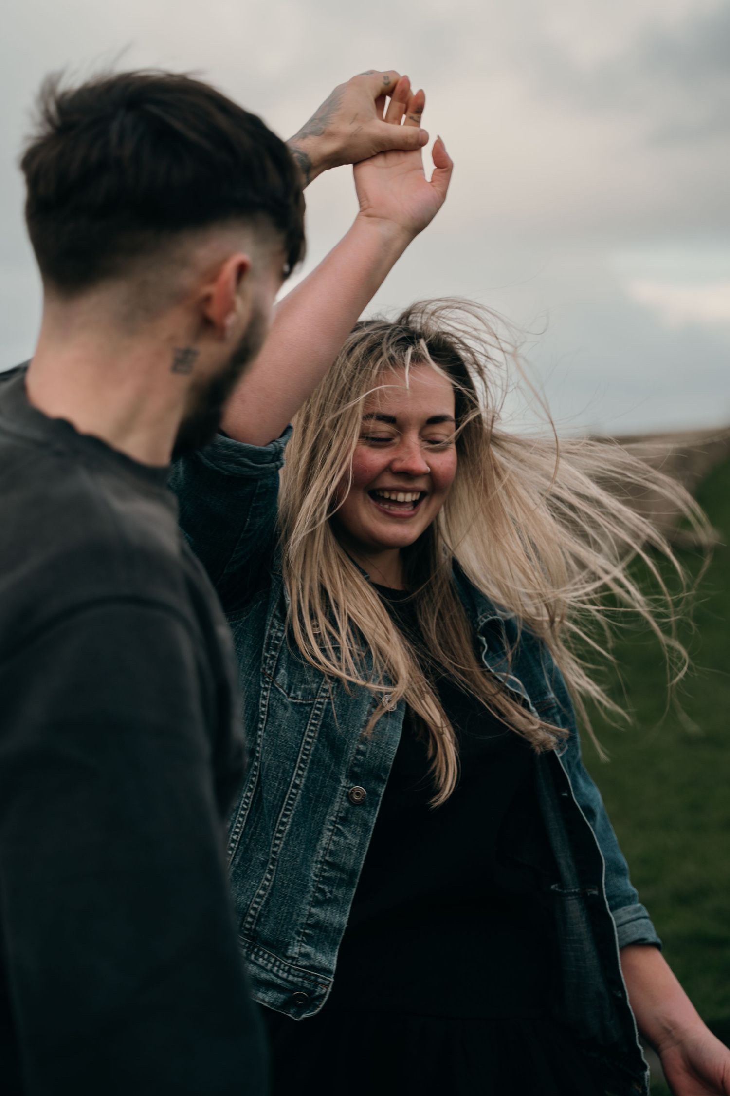 Joyful blonde woman spinning and laughing outdoors with her partner on a cloudy day.