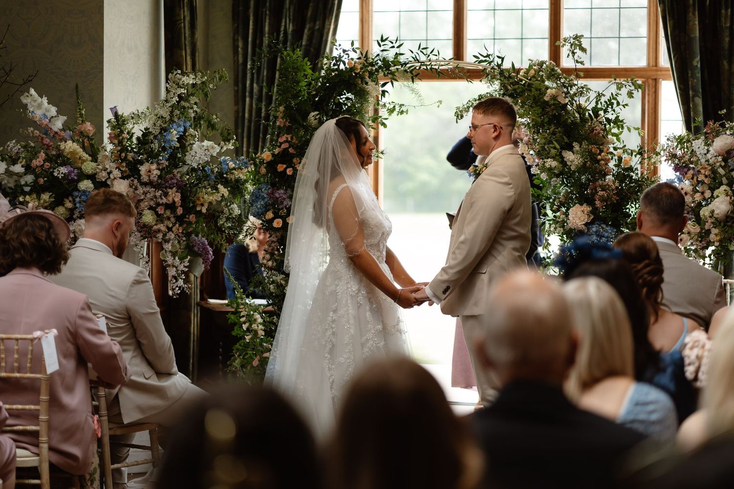 Bride and groom exchange vows at floral arch altar during indoor wedding ceremony with seated guests.