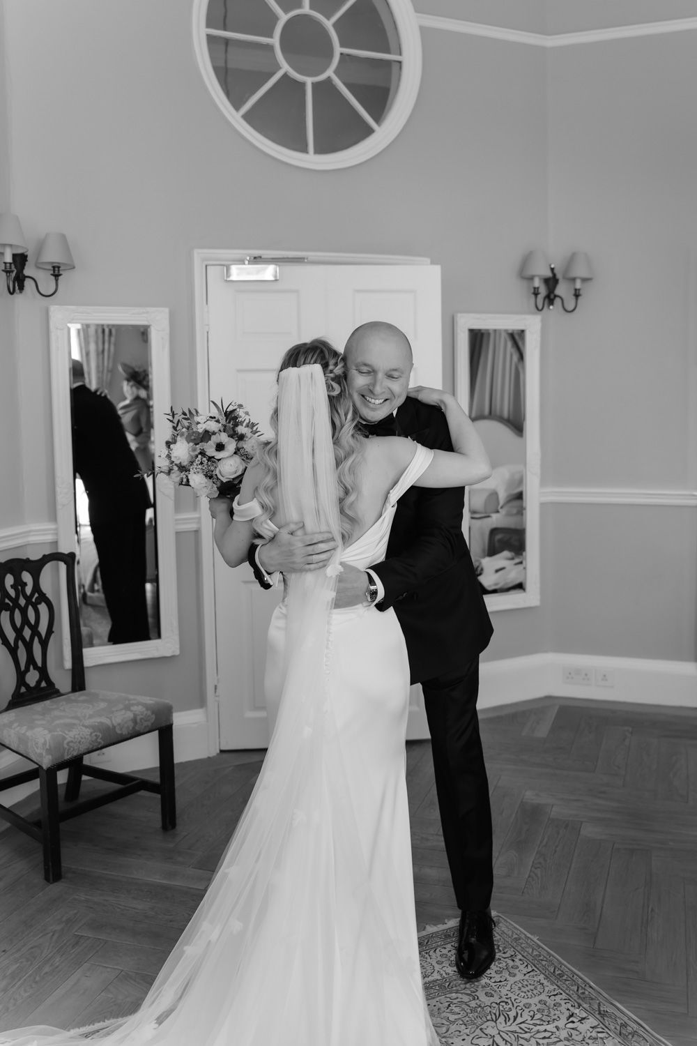 Groom embracing bride in stunning off-shoulder gown during emotional first look in elegant room.
