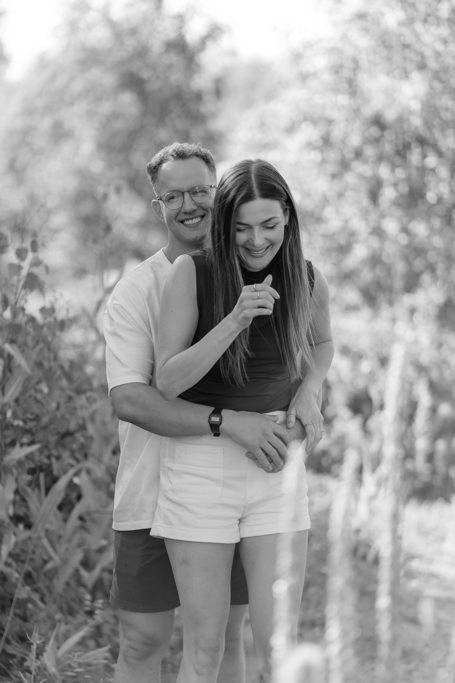 Black and white photo of a smiling couple embracing outdoors, man hugging woman from behind in a lush garden setting.