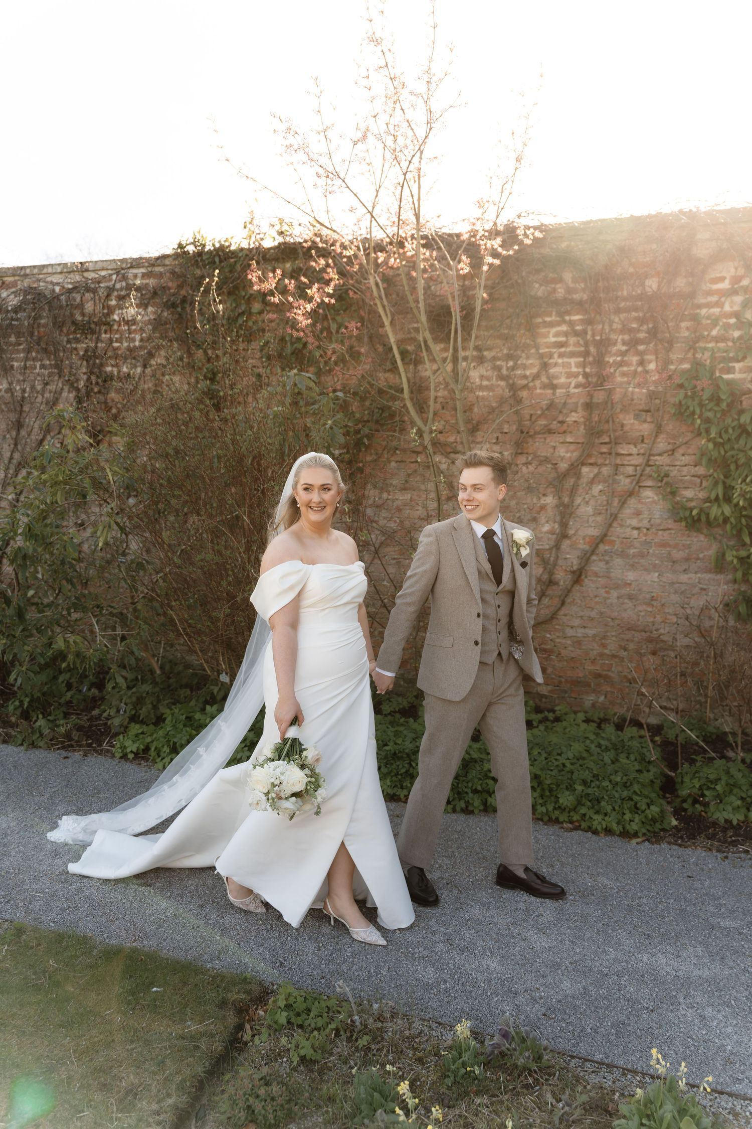 Bride in white off-shoulder gown and groom in grey suit walk hand-in-hand along path at golden hour.
