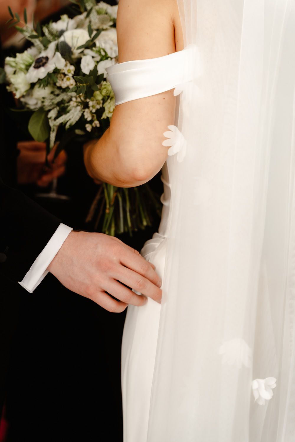 Close-up of groom's hand holding bride's white veil with floral appliques, bride holding white bouquet.