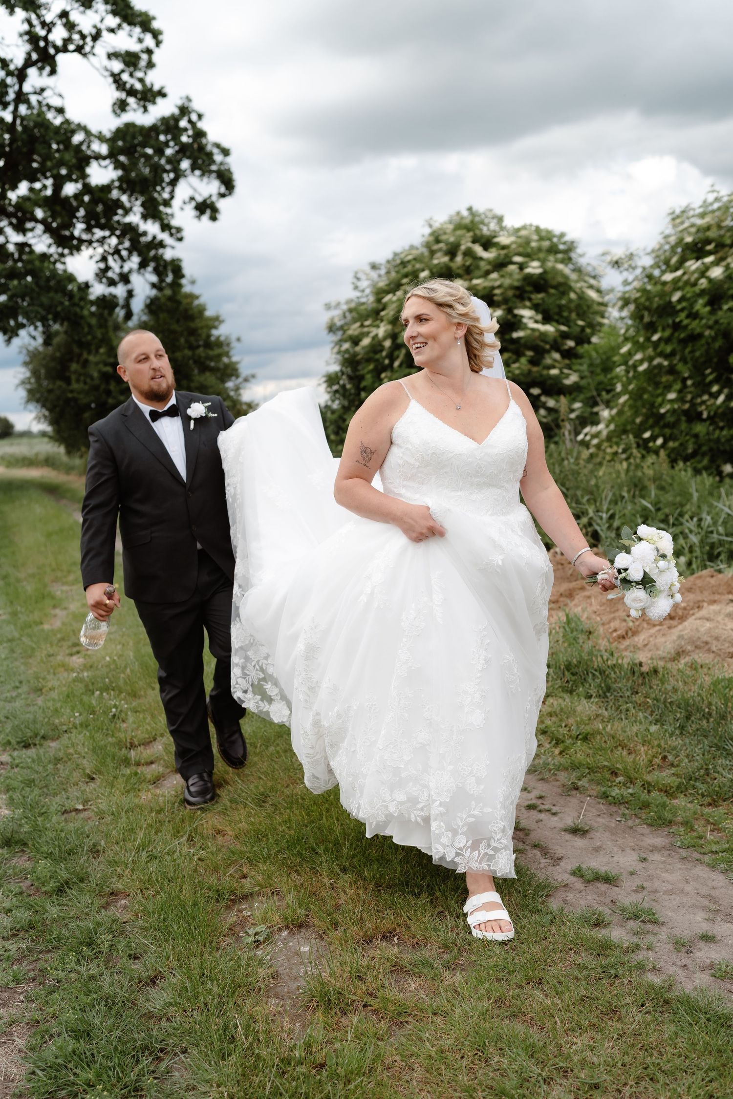 Smiling bride in white lace gown walks with groom in black tuxedo along a rural path on their wedding day.