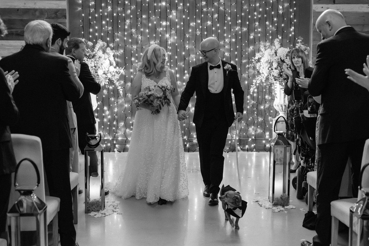 Black and white photo of bride and groom walking down aisle with dog at wedding ceremony.