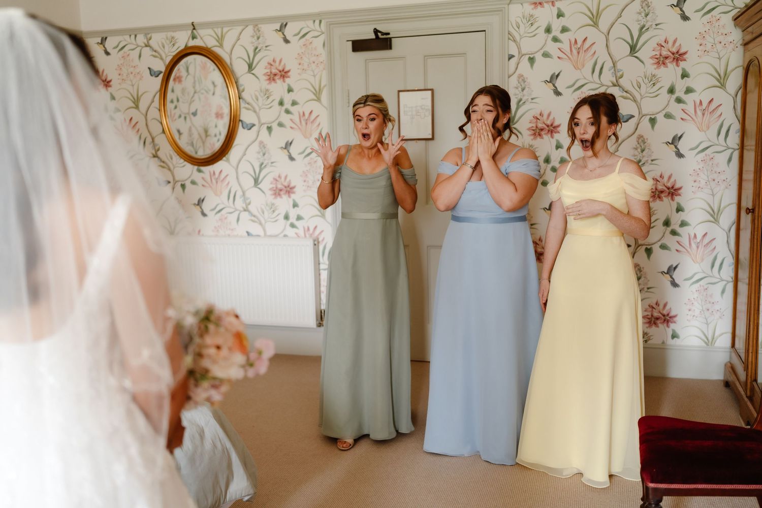 Three bridesmaids in pastel gowns react with joy and tears seeing the bride in her wedding dress and veil.
