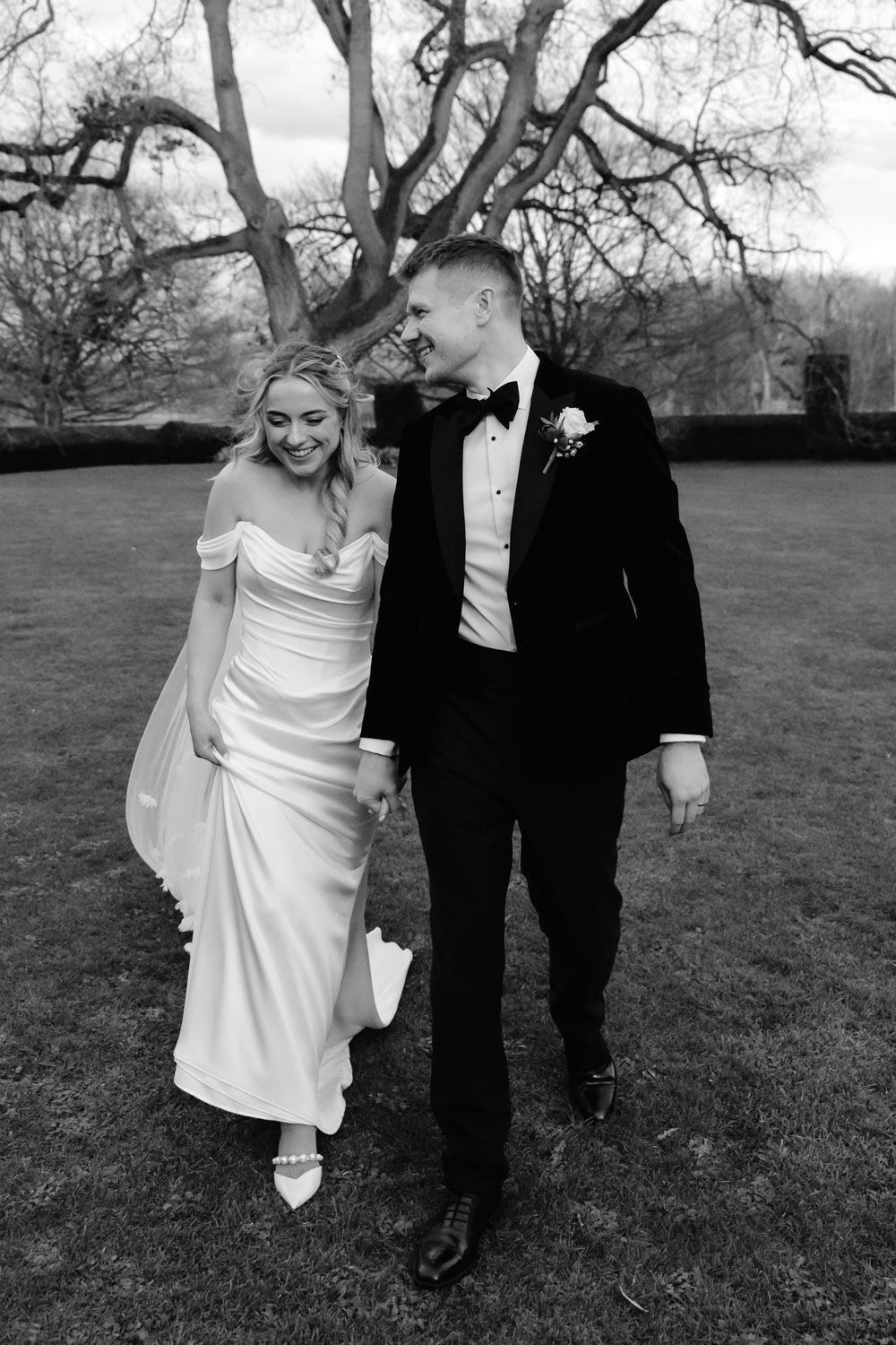 Black and white photo of a smiling bride and groom walking hand in hand across a lawn on their wedding day.