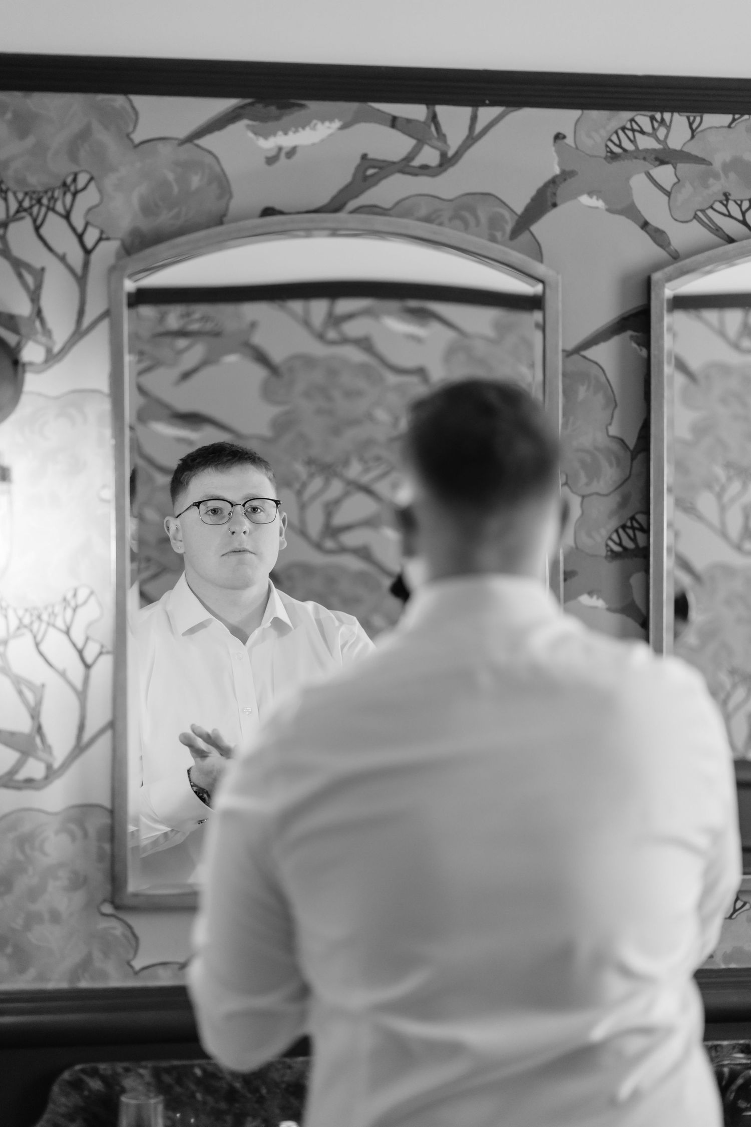 Black and white photo of a man in a white shirt adjusting his outfit while looking at his reflection in an ornate mirror.