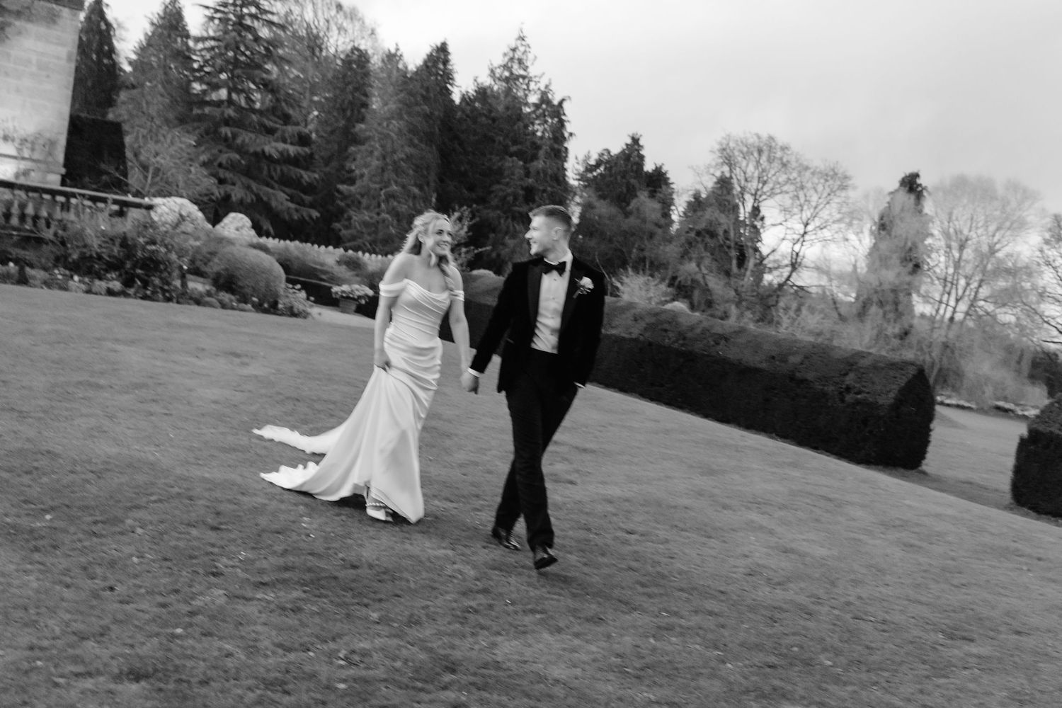 Black and white photo of bride and groom holding hands walking across a manicured lawn at their wedding venue.