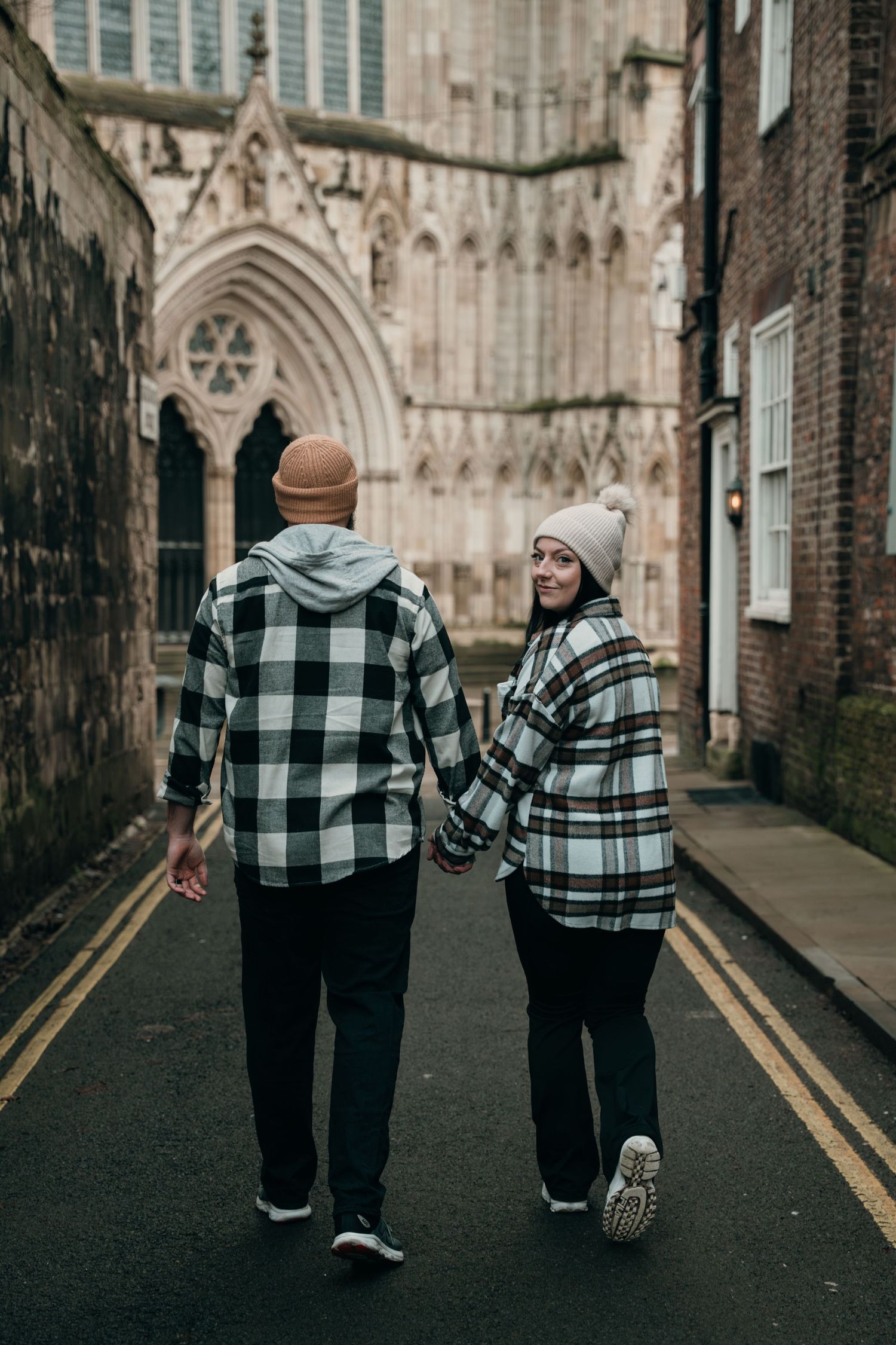 A couple in plaid shirts walk hand in hand toward a stunning Gothic cathedral on a quiet street.