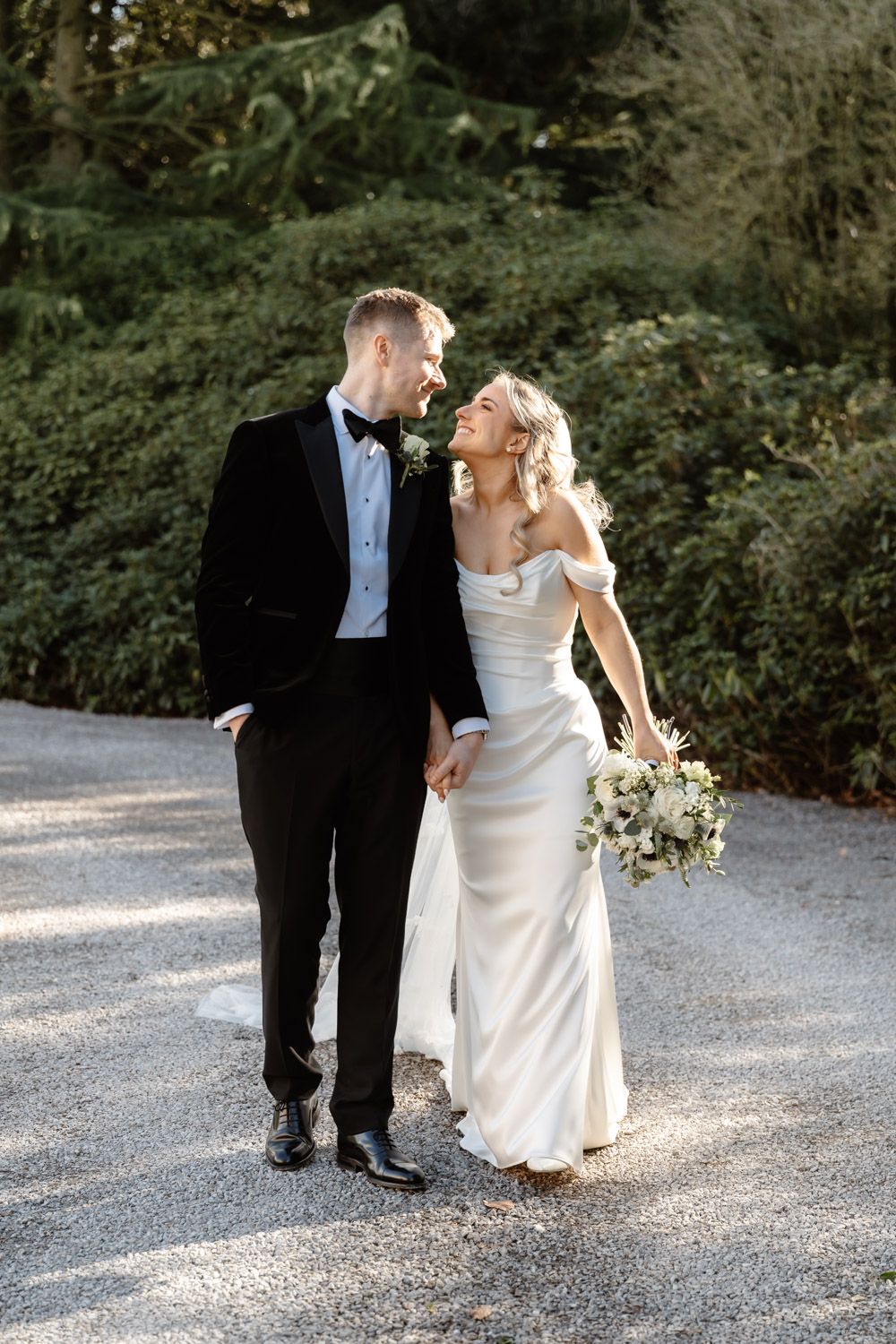 A smiling bride in an off-shoulder white gown holds a bouquet while walking with a groom in a black tuxedo outdoors.