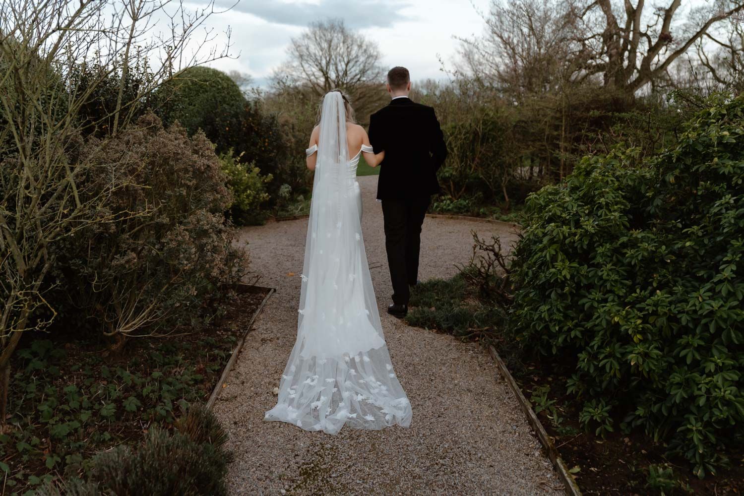 Bride with long lace cathedral veil walks garden path with groom from behind.
