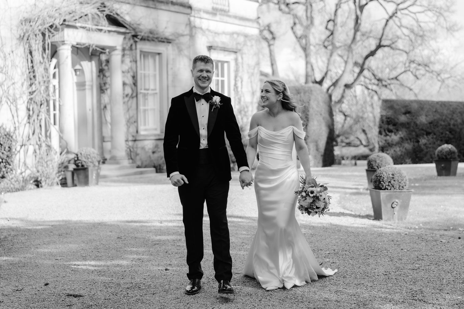 Bride and groom smiling while walking outside a historic manor in black and white.